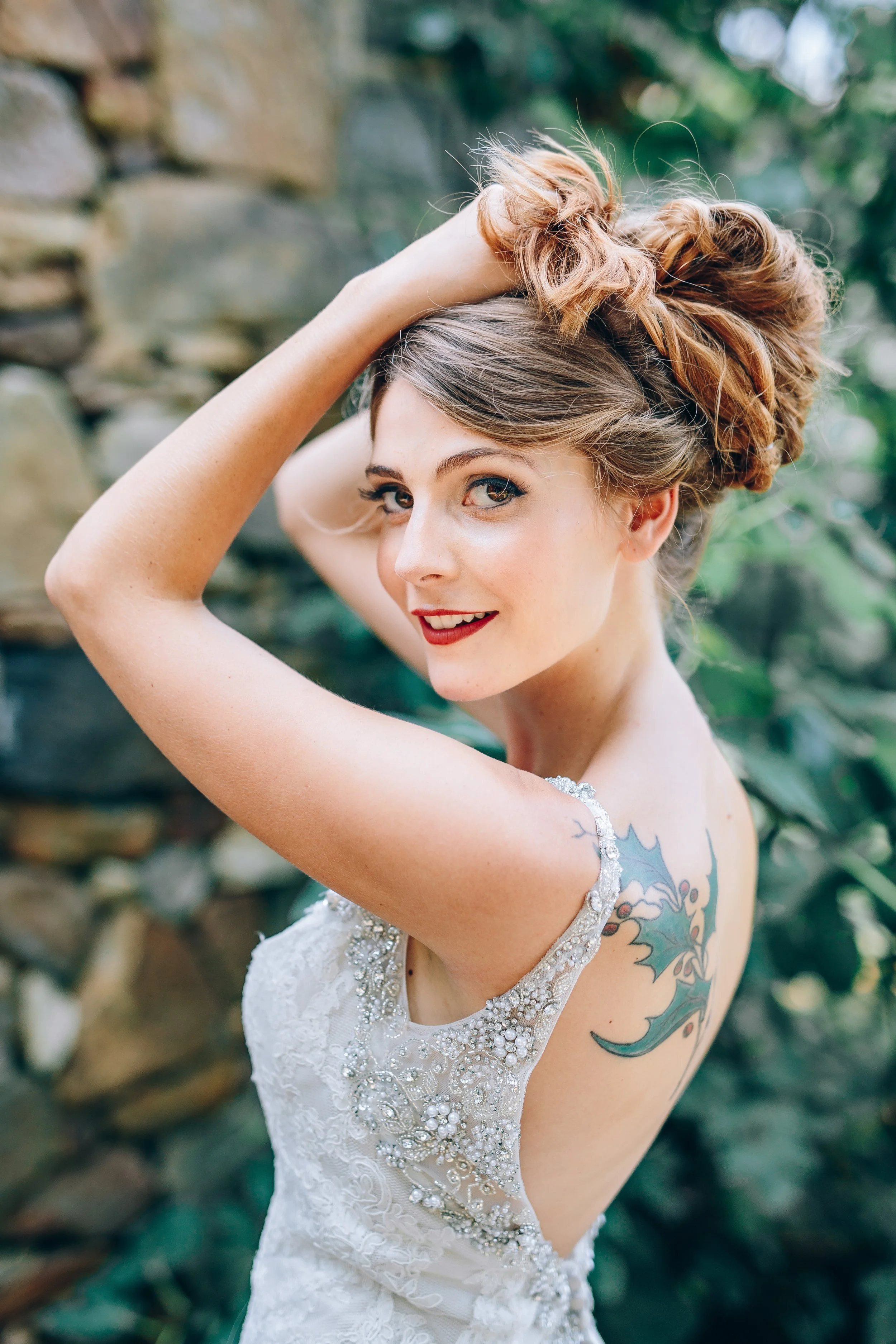 A bride with a tattoo on her back, wearing a wedding dress with embellished details, smiling with her hand on her head, outdoors with greenery and rocks in the background. Atlanta Georgia.  Wedding photography by Sowing Clover Photography.