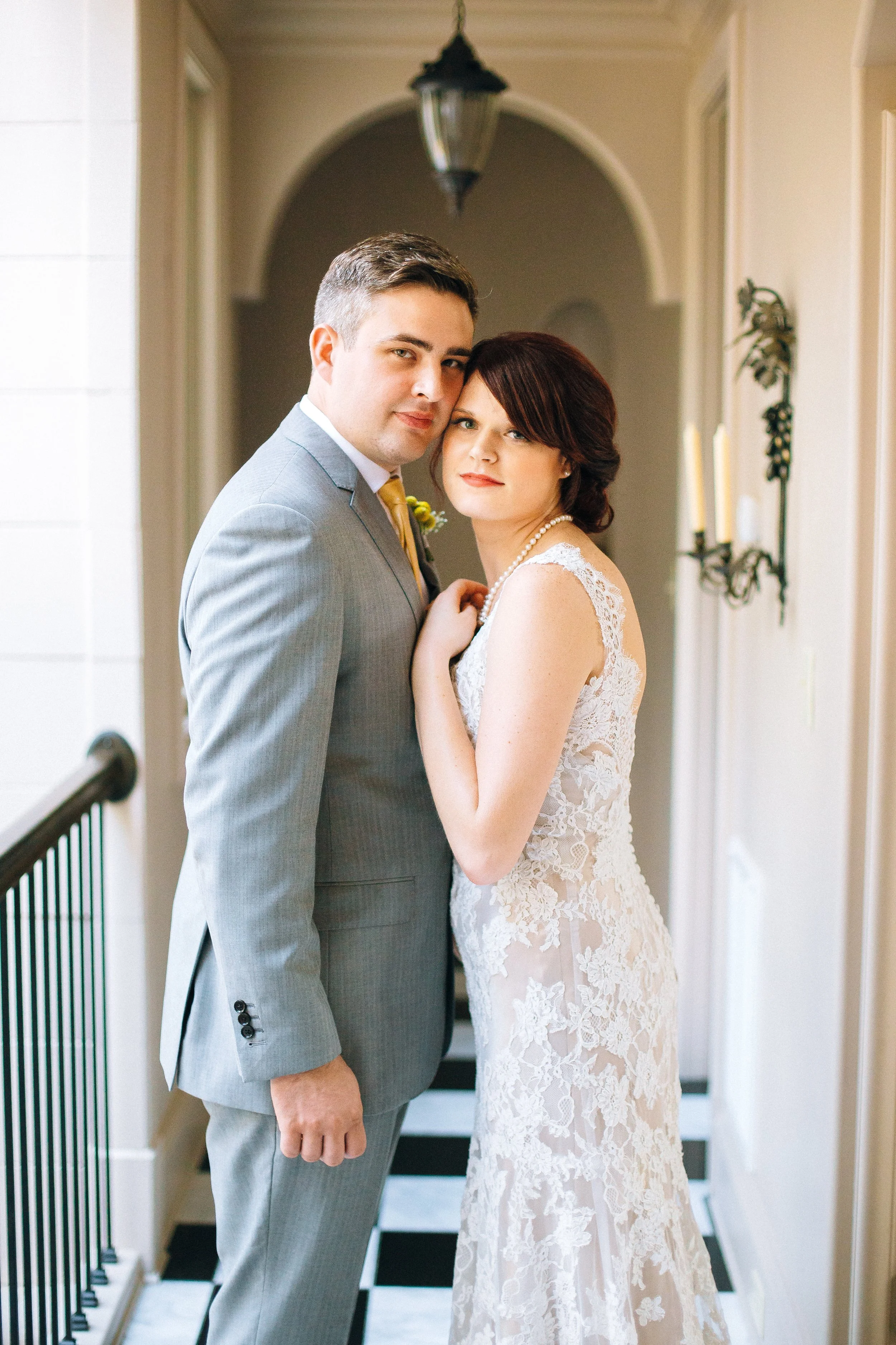 A bride and groom in wedding attire standing close together indoors, with the bride in a lace wedding dress and the groom in a gray suit in Atlanta. Wedding Photography in by Sowing Clover Photography. 