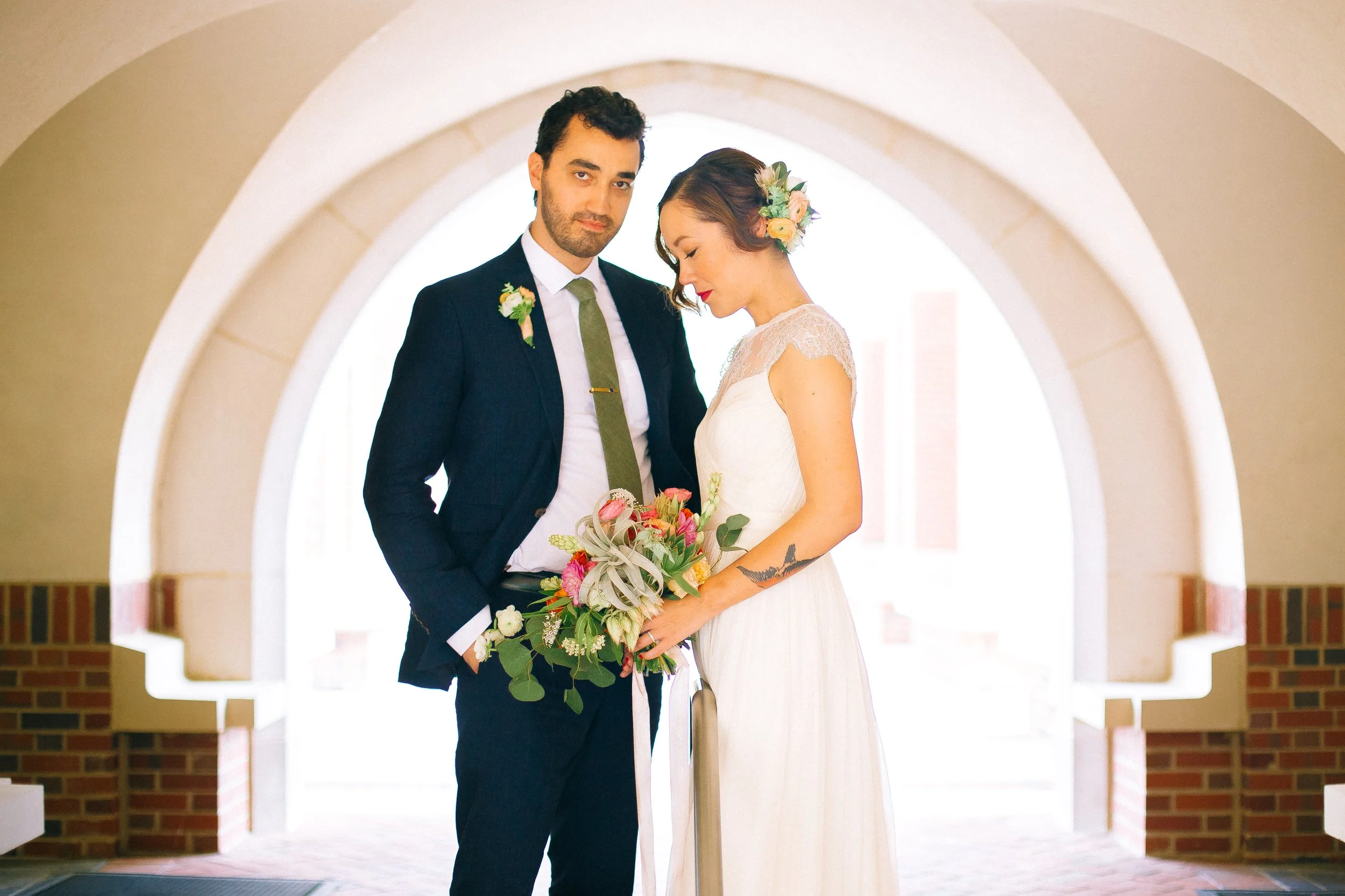 A bride and groom standing together in front of a large bright archway, with the bride holding a colorful bouquet and the groom dressed in a dark suit with a boutonniere in Decatur, GA. Wedding Photography by Sowing Clover Photography.