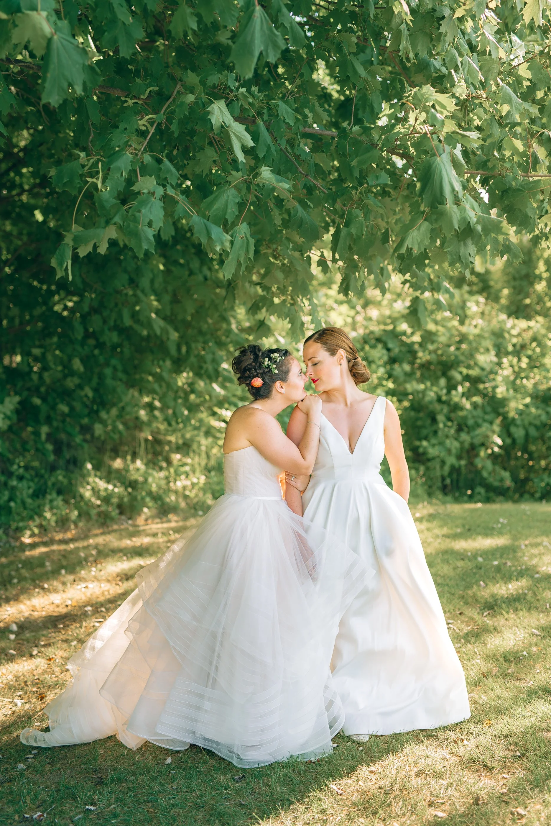 Two women in wedding dresses standing under a tree, sharing a tender moment outdoors. Wedding Photography by Sowing Clover Photography.