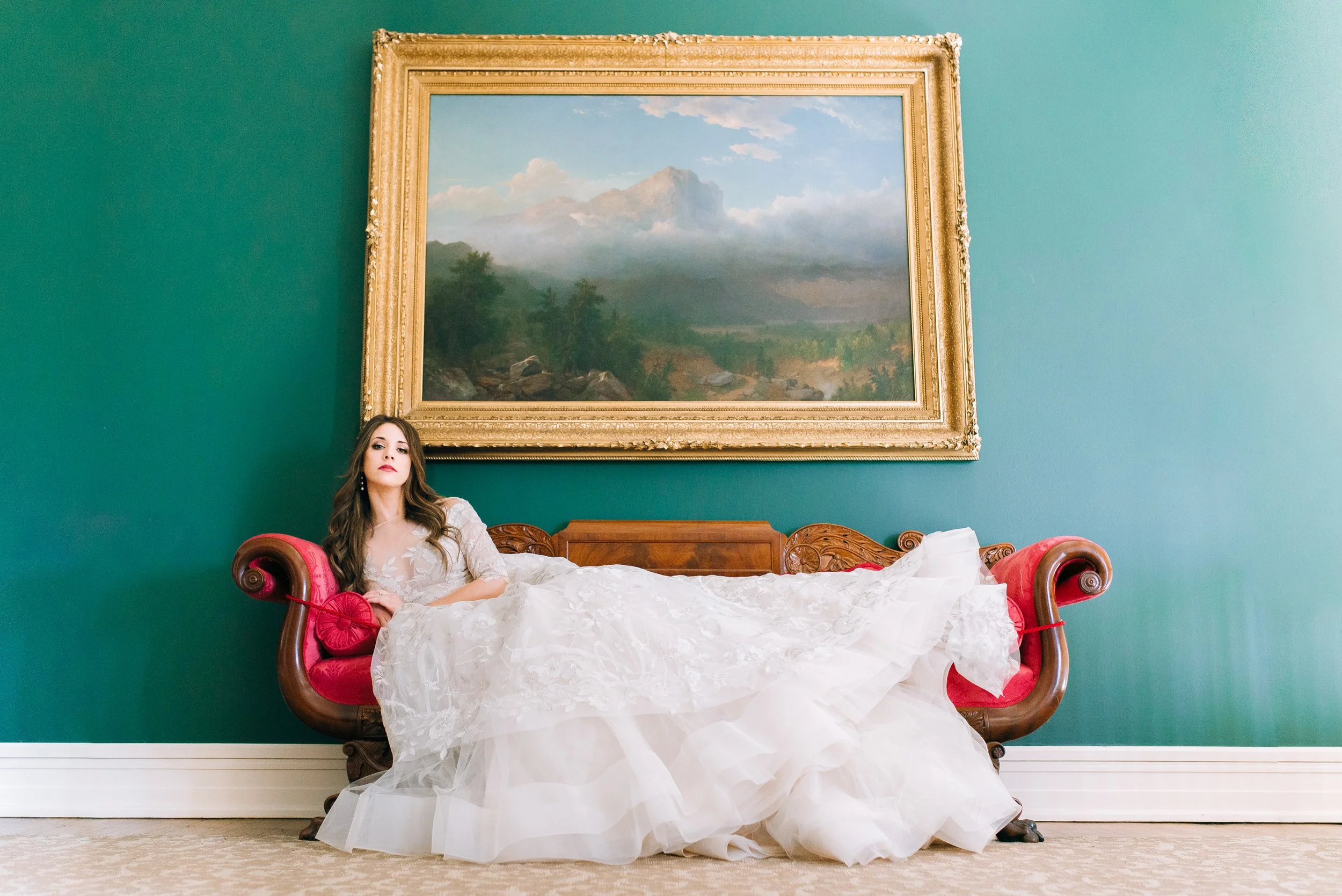 A bride in a white wedding dress is reclining on a vintage wooden sofa with red upholstery in front of a large landscape painting in a gold frame at the Hunter Museum of American Art. Wedding Photography by Sowing Clover Photography.