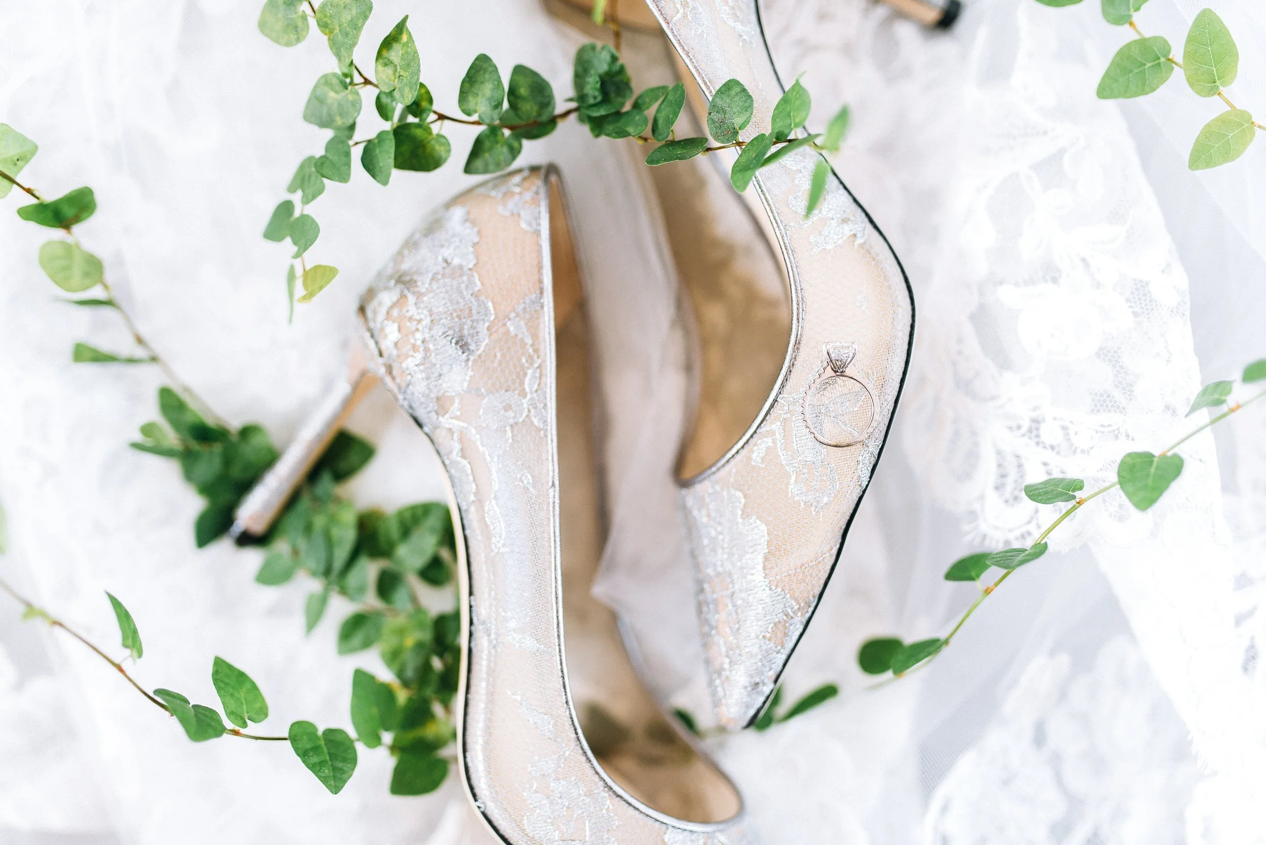 Elegant Jimmy Choose lace high-heeled wedding shoes with black piping, adorned with a diamond engagement ring placed on one of the shoes, with green leafy vines on a white lace fabric background. The Estate Atlanta. Sowing Clover Wedding Photography.