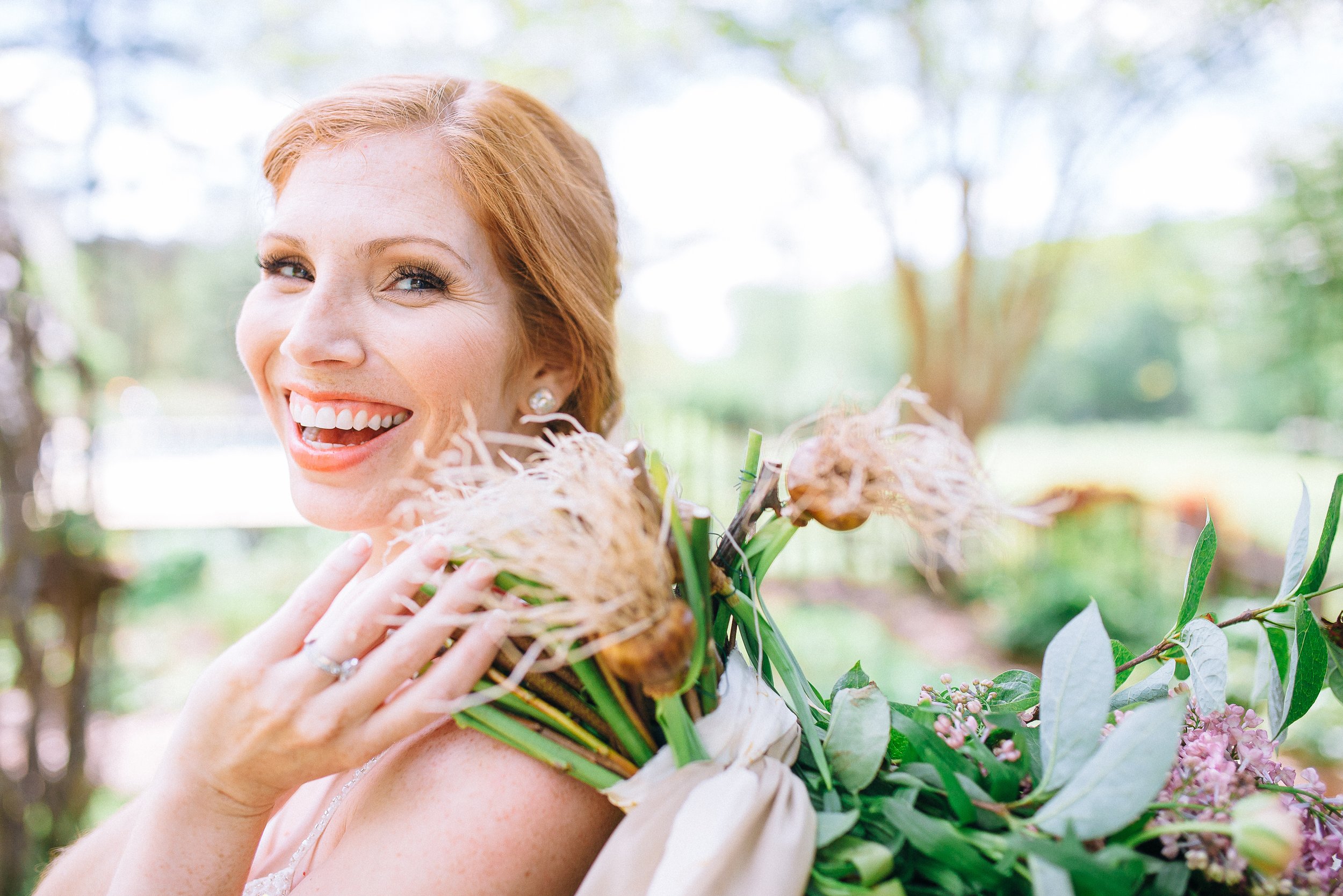 A bride with red hair and earrings smiling while holding a large bouquet of flowers outdoors on a sunny day at Glen Ella Springs.  Wedding Photography by Sowing Clover Photography.