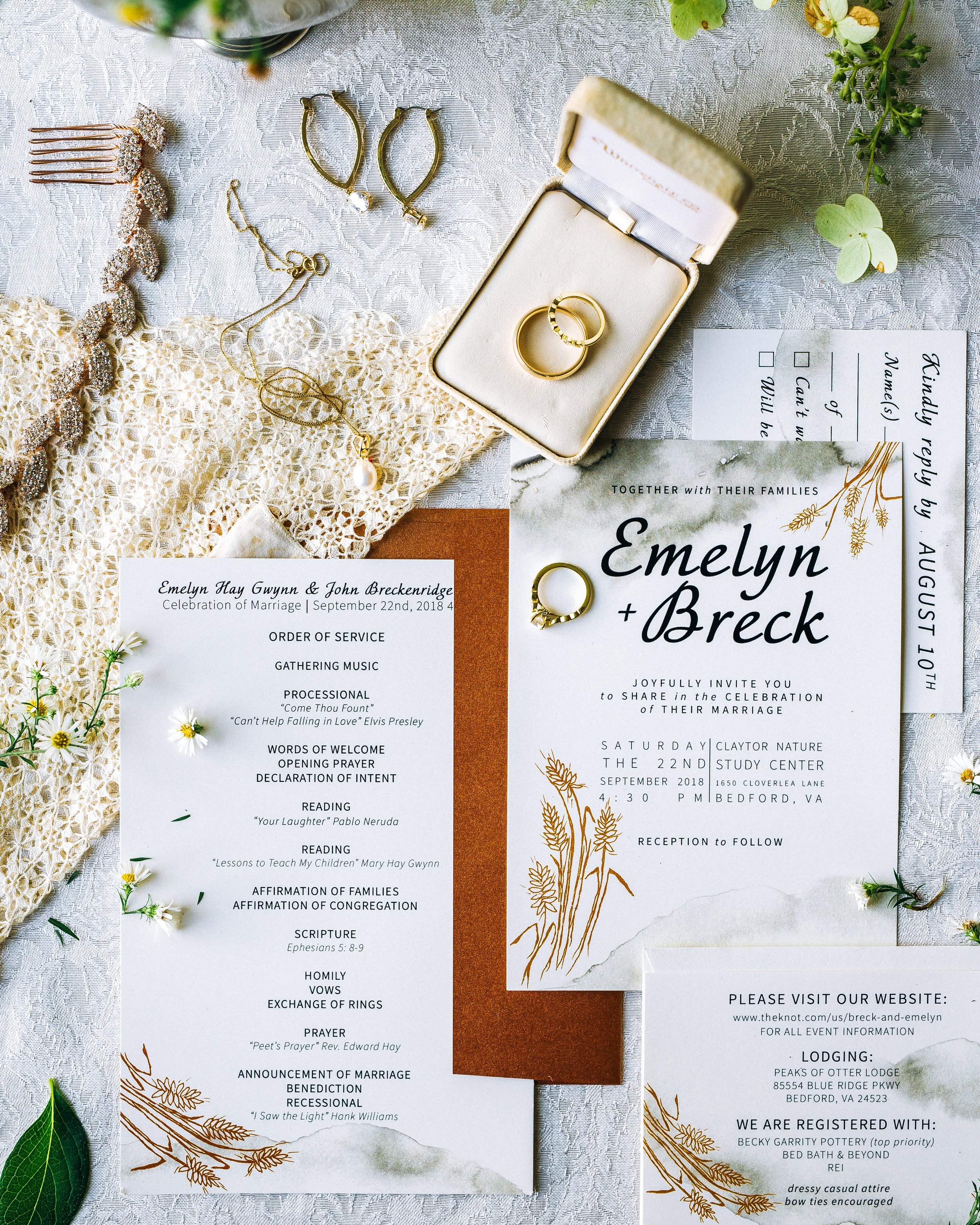 Wedding invitations, rings, jewelry, and a ring box arranged on a lace and fabric surface, accented with small flowers and greenery. Wedding Photography by Sowing Clover Photography.