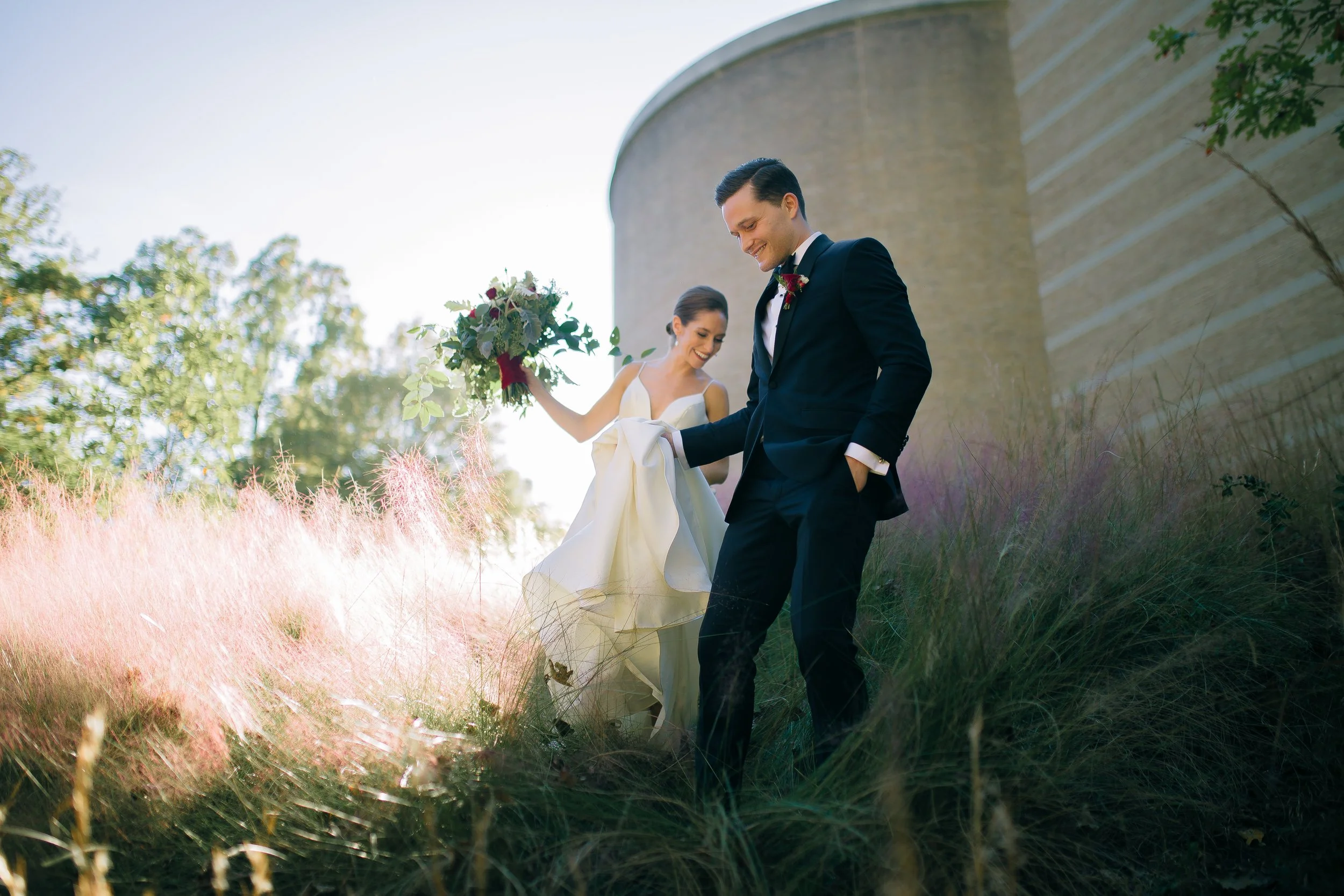 A bride and groom in wedding attire outdoors, standing on pink and purple grass near a large concrete wall. The bride is holding a bouquet and smiling at Fernbank Science Center. Wedding Photography by Sowing Clover Photography.