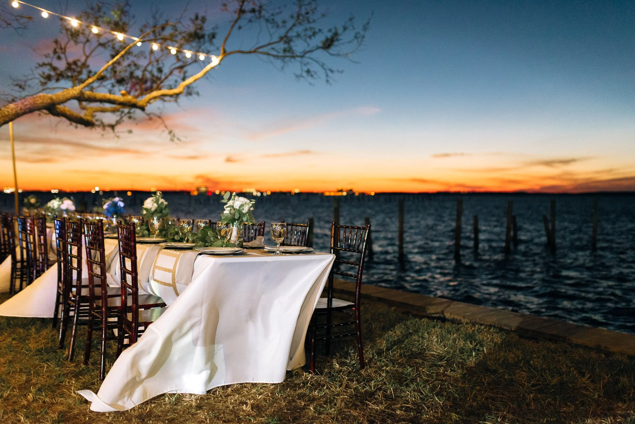 A beautifully decorated outdoor dining table set beside a body of water at sunset with string lights hanging from a nearby tree in Florida for a wedding. Wedding Photography by Sowing Clover Photography.