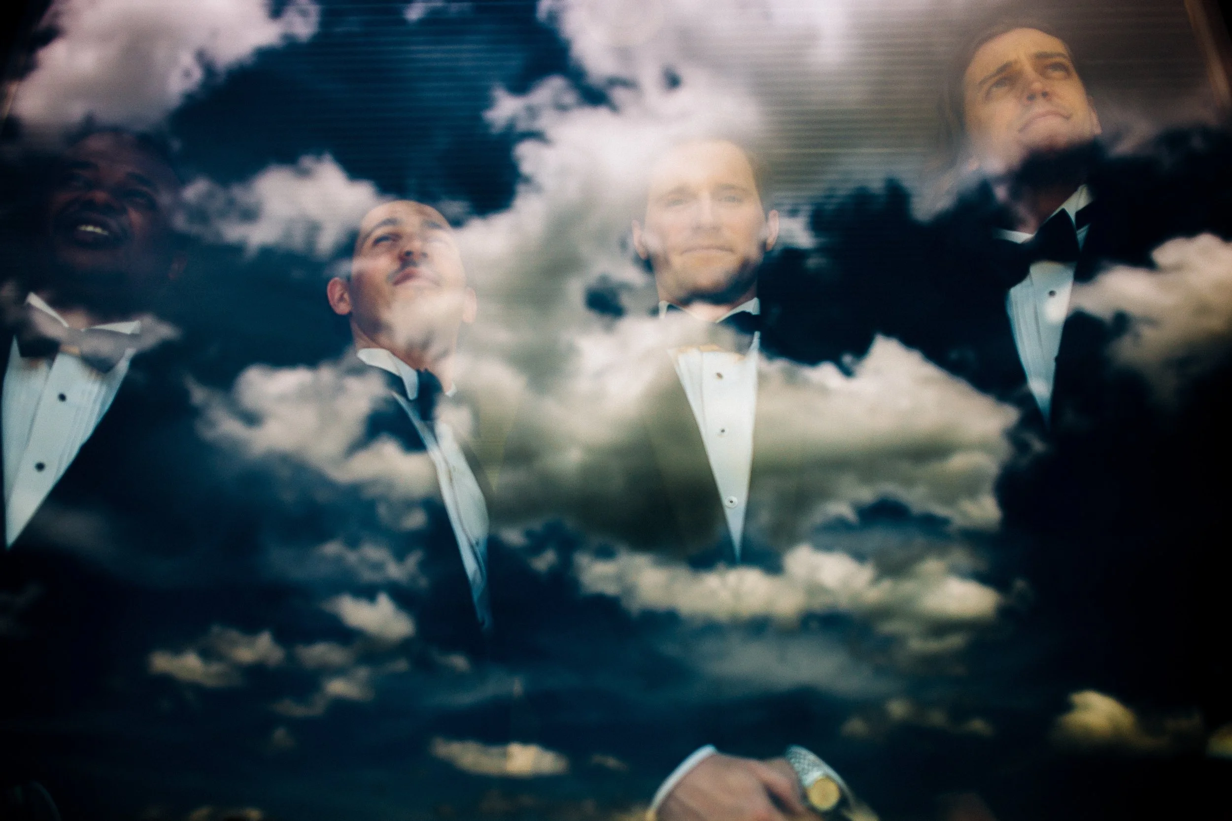 Reflection of four men dressed in tuxedos looking up, seen through a window with clouds reflecting on the glass on the groom's wedding day for his wedding photography at The Farm wedding venue in north Georgia 
