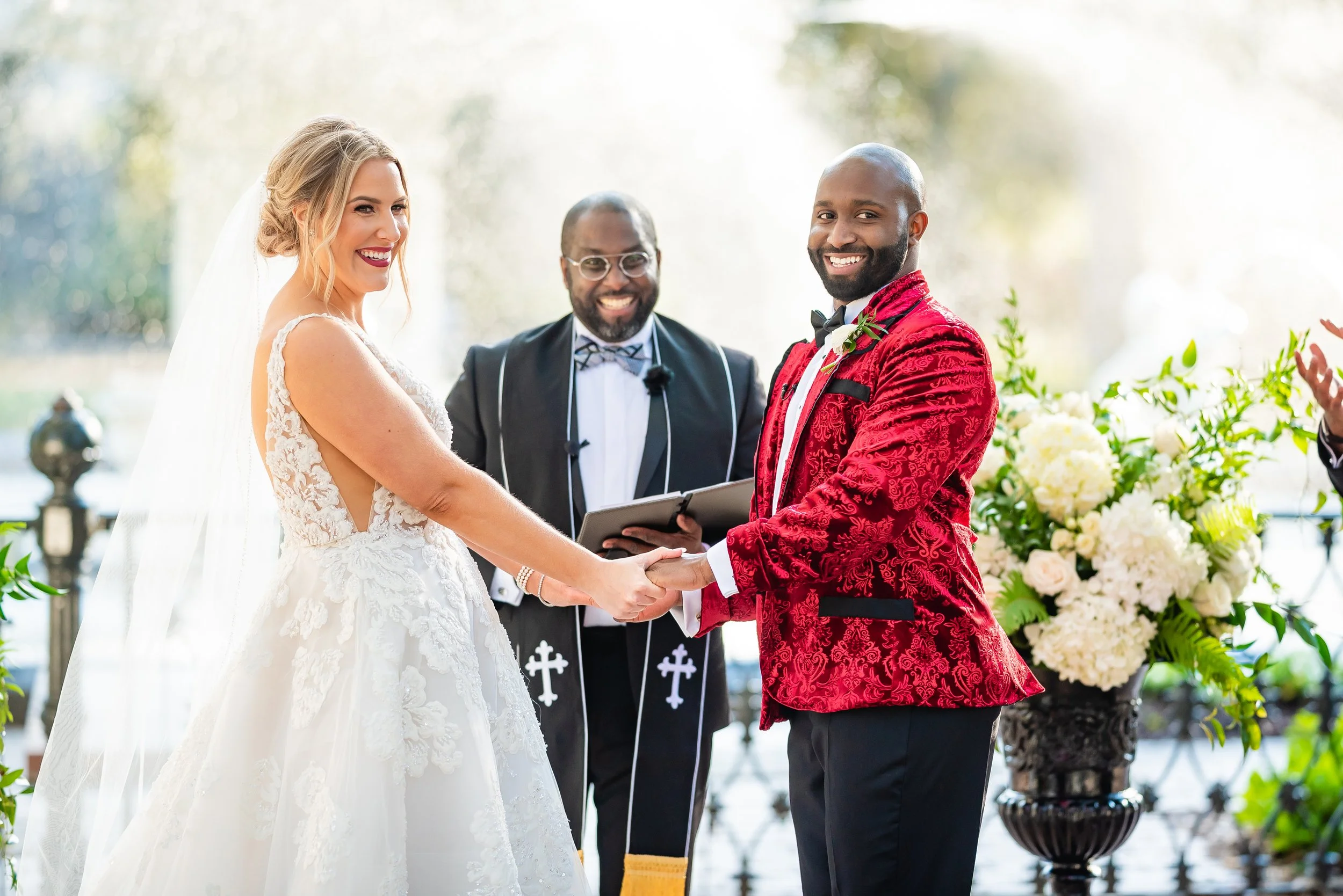 A wedding ceremony with a bride and groom holding hands and smiling, officiant behind them, outdoors with flowers and greenery in front of the fountain at Forsyth Park in Savannah, Georgia. Wedding Photography by Sowing Clover Photography.