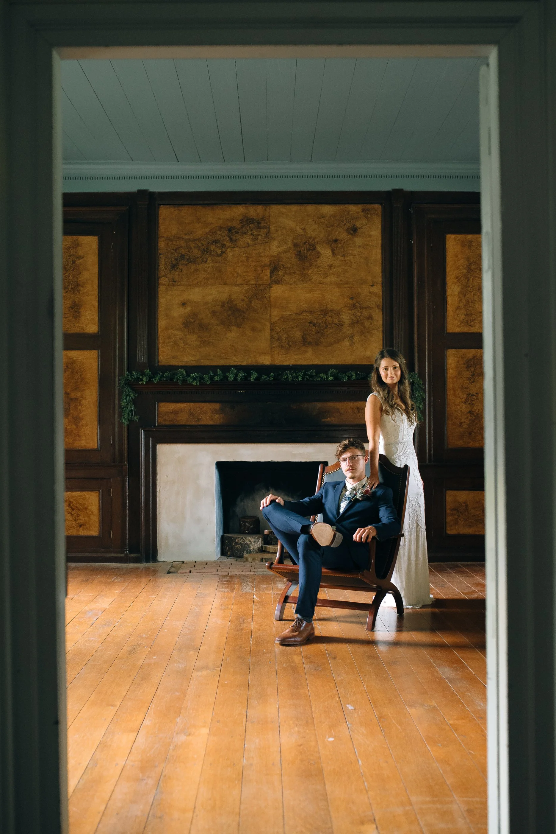 A groom in a navy blue suit with glasses, sitting on a vintage wooden chair, and a bride in a white dress standing behind him at Historic Rock Castle in Hendersonville. Wedding Photography by Sowing Clover Photography.