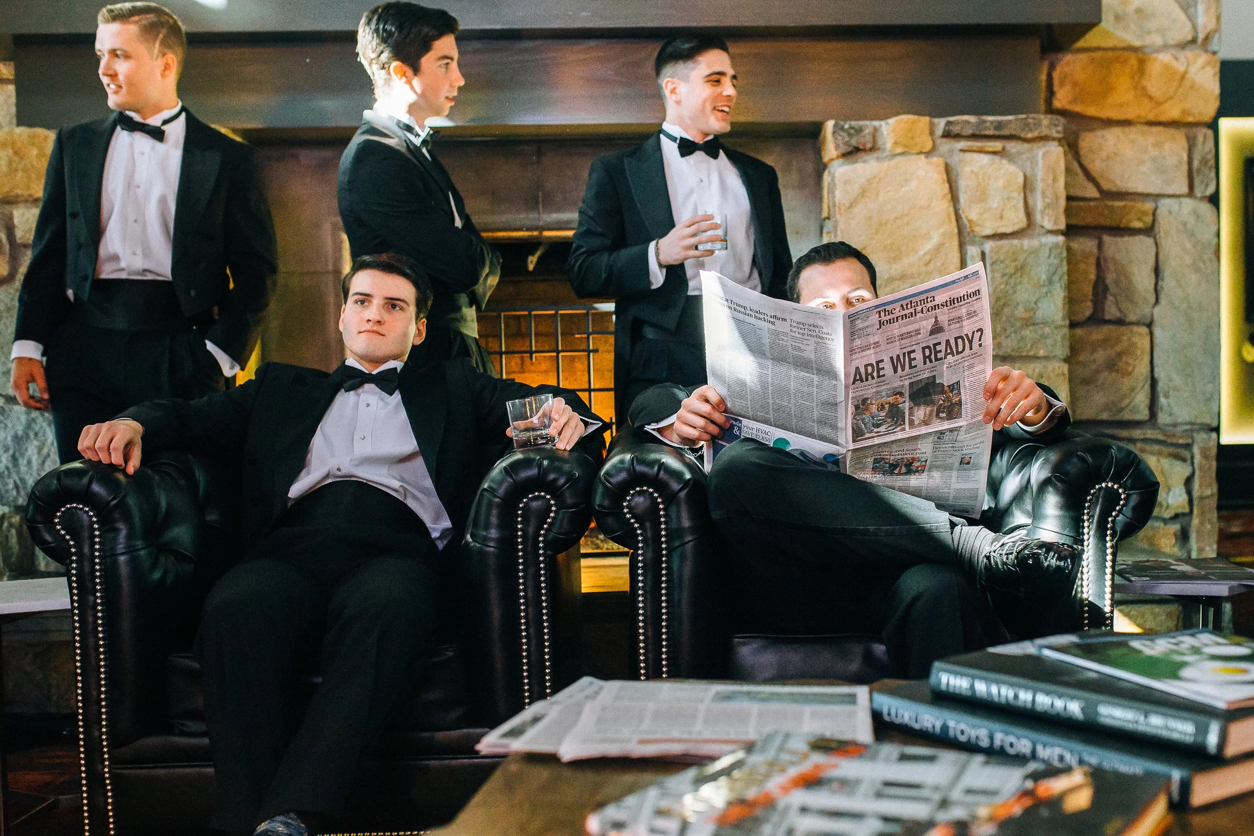 Four men dressed in formal tuxedos in a lounge area. Two seated on a black leather couch, one reading a newspaper and the other with a drink, and two standing behind them, engaged in conversation, in front of a stone fireplace.