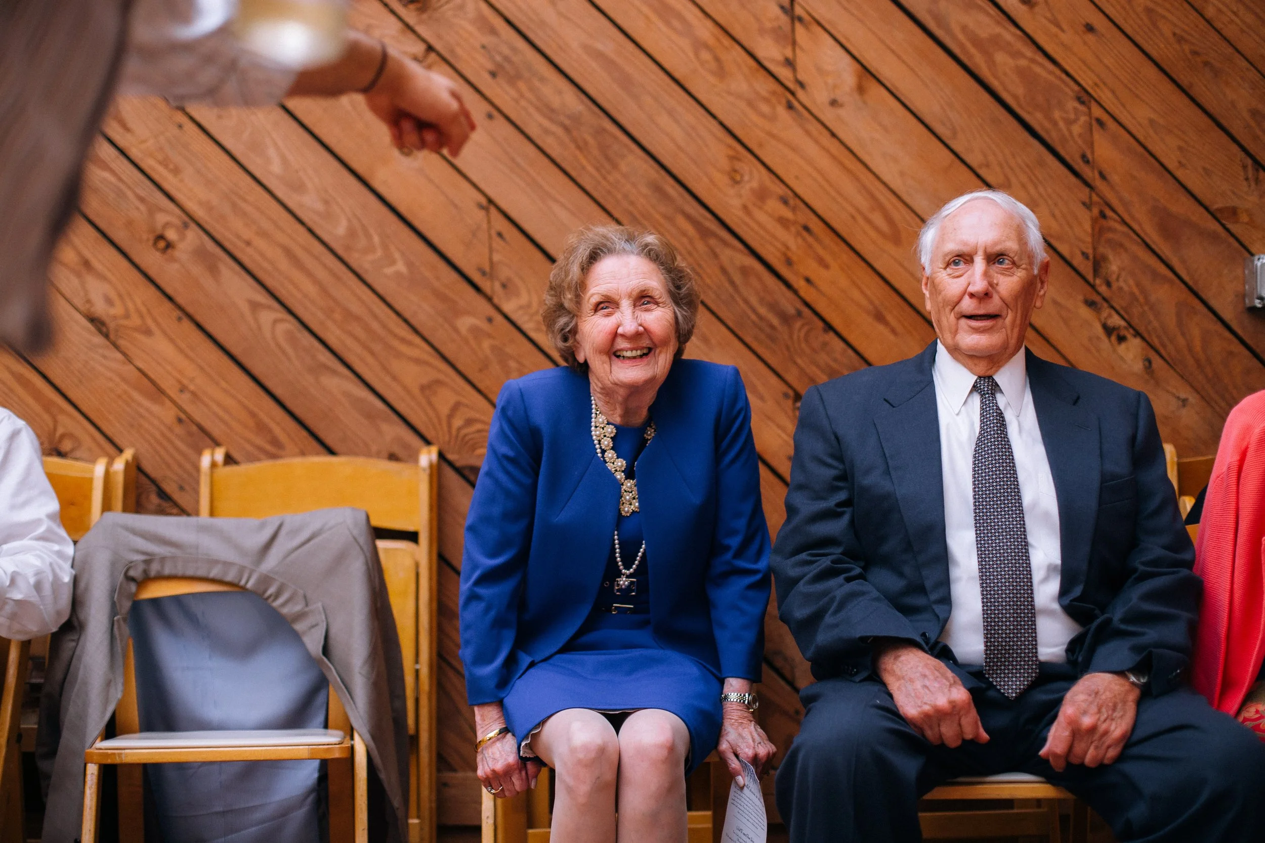 An elderly woman and man sitting in a row of chairs, smiling and looking at a person who is gesturing or speaking in front of them. The woman is wearing a blue dress and blazer. Nashville Farm Wedding Wedding photography by Sowing Clover Photography.