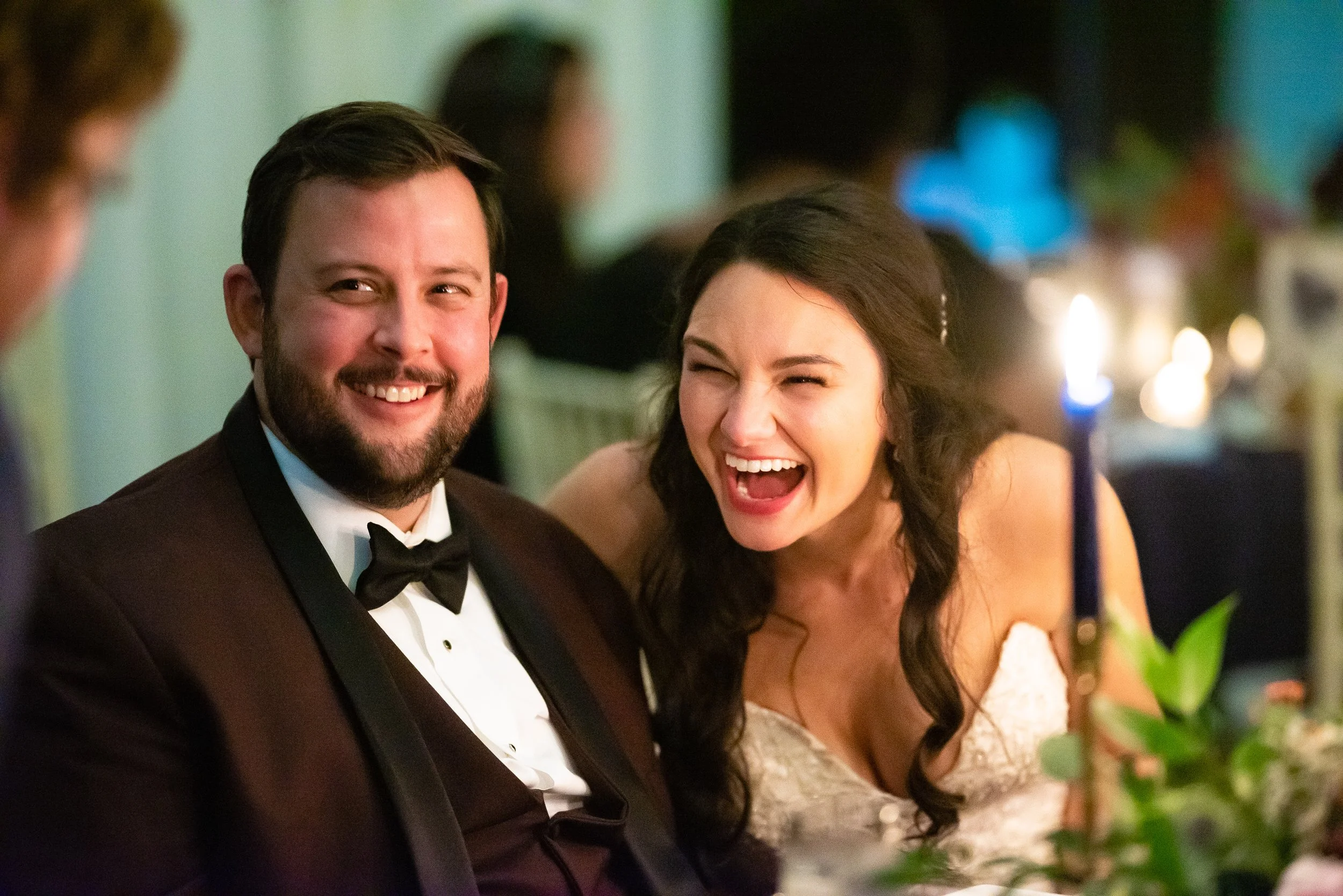 A groom in a tuxedo and a bride in a white dress laughing at a formal event table with a candle and floral arrangement at The Atrium in Norcross GA Wedding photography by Sowing Clover Photography