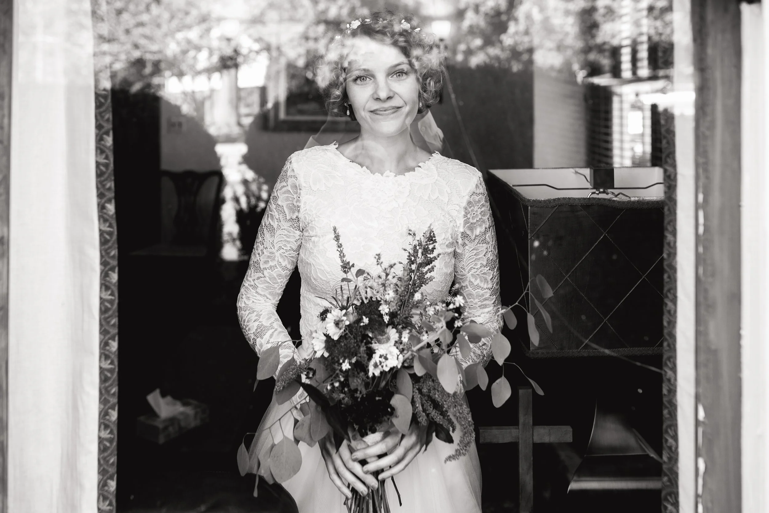 A bride in a lace wedding dress holding a bouquet of flowers, standing behind a window in Nashville TN Wedding photography by Sowing Clover Photography