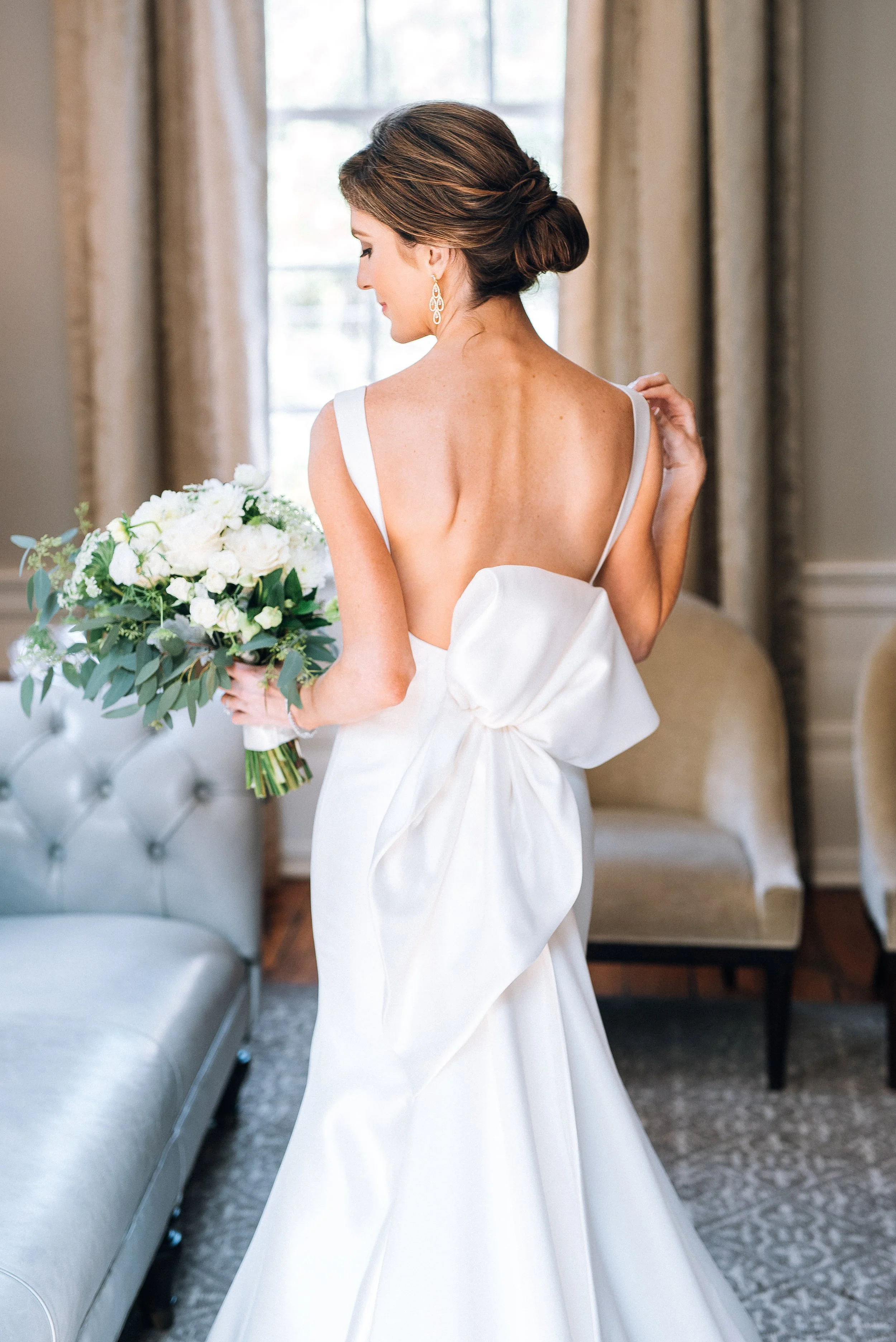 Bride in a white wedding dress holding a bouquet, viewed from the back, inside a room with beige curtains and vintage chairs at The Estate on Piedmont Road in Atlanta, GA. Wedding Photography by Sowing Clover Photography.