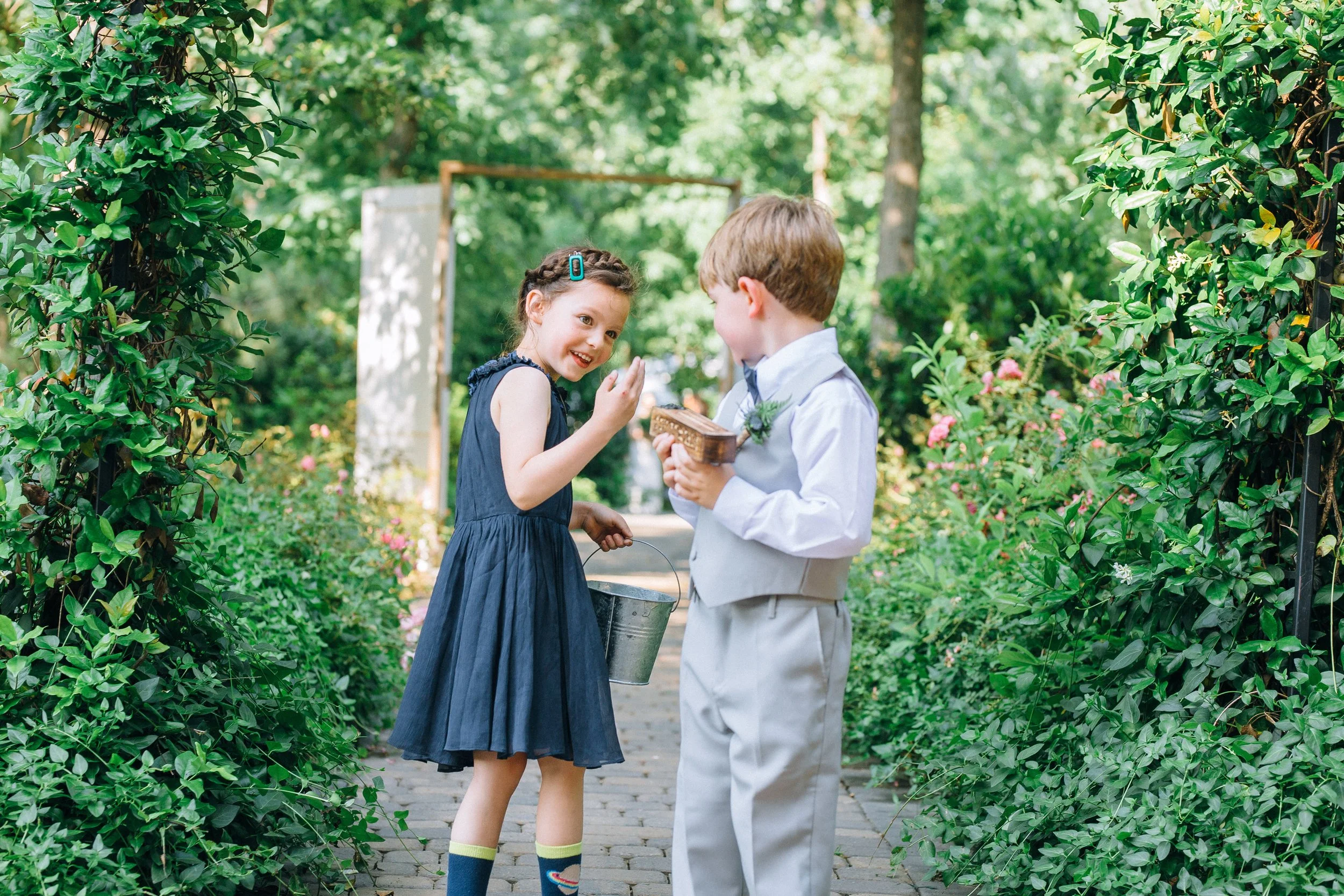 A flower girl in a blue dress and a young ring bearer in a light-colored suit exchanging gifts or tokens in a lush green garden walkway before a wedding ceremony in Nashville TN Wedding photography by Sowing Clover Photography.