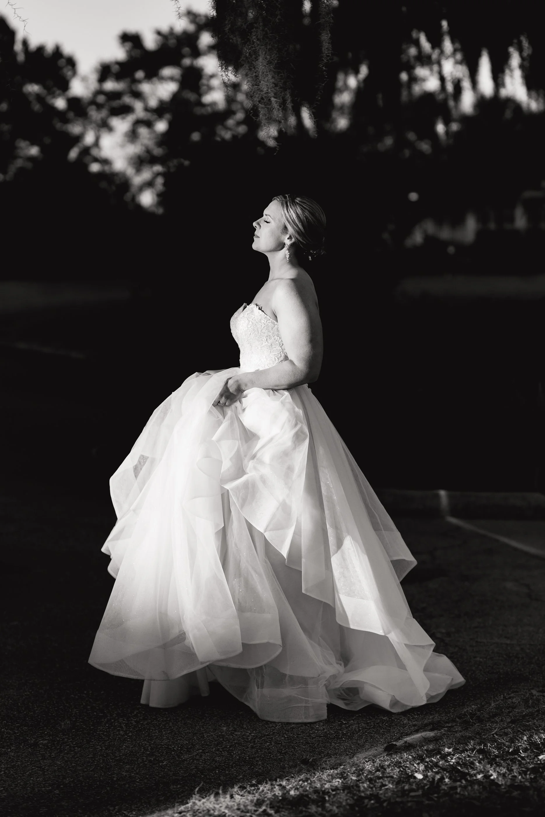 A bride in a wedding dress standing outdoors at dusk, her eyes closed and face turned upwards, holding her gown, with trees and a dark sky in the background. Orlando Country Club Wedding. Wedding photography by Sowing Clover Photography.