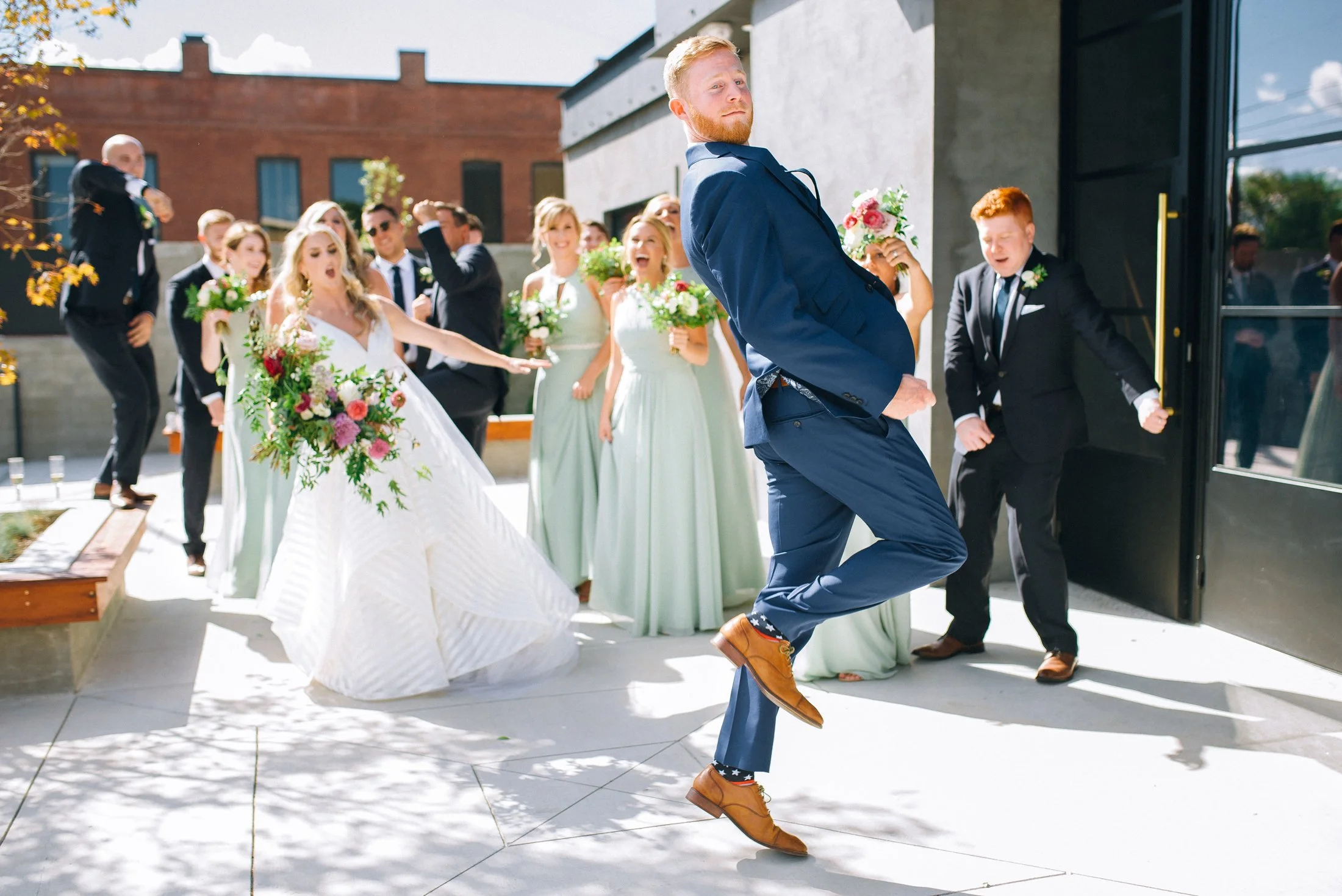 A groom in a blue suit is dancing in front of a group of bridesmaids and groomsmen at a wedding celebration outside of Clementine Hall in Nashville, TN.  Wedding Photography by Sowing Clover Photography.