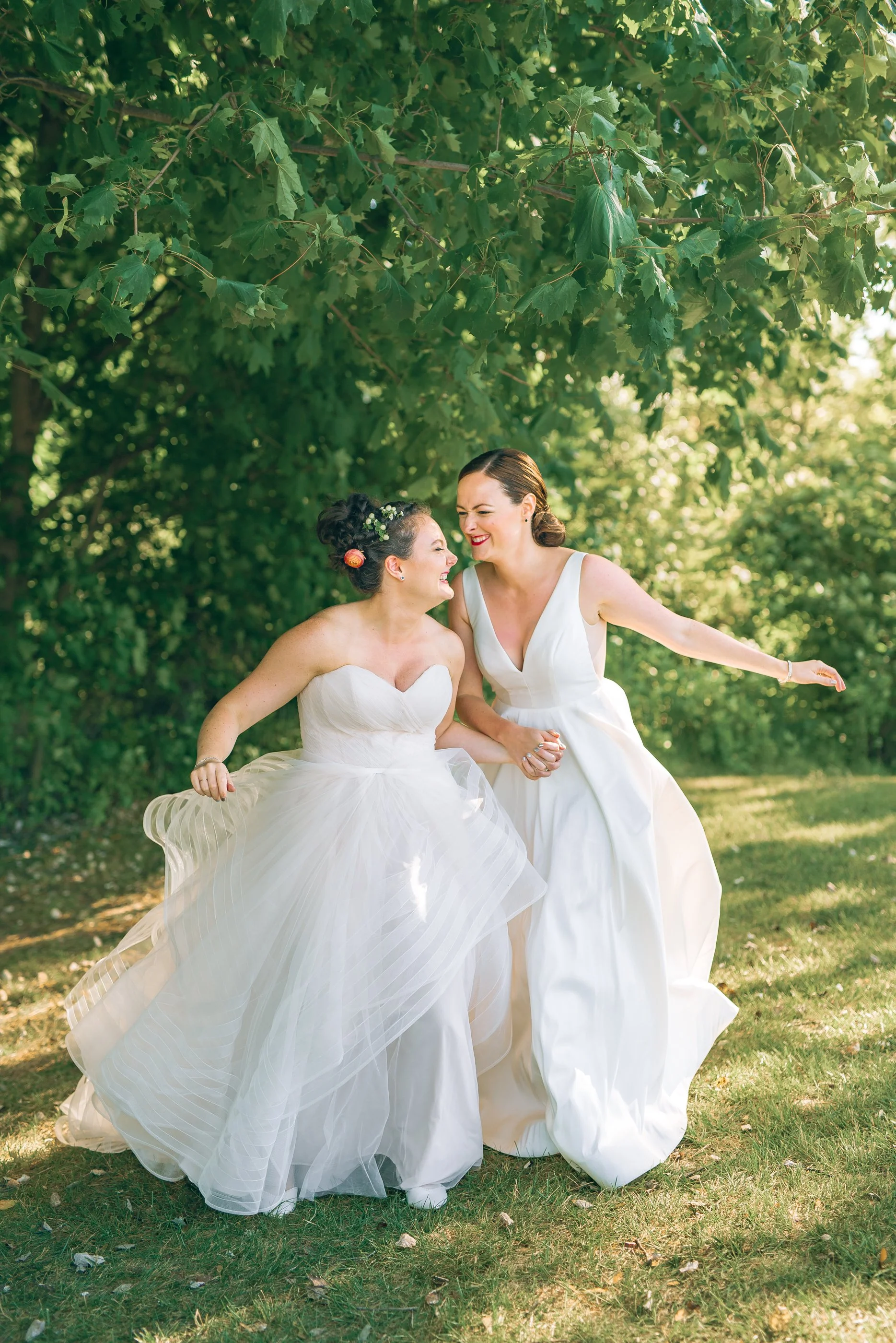 Two brides in white wedding dresses laughing and holding hands outdoors with green leaves overhead for a lesbian wedding torch lake Michigan Wedding photography by Sowing Clover Photography