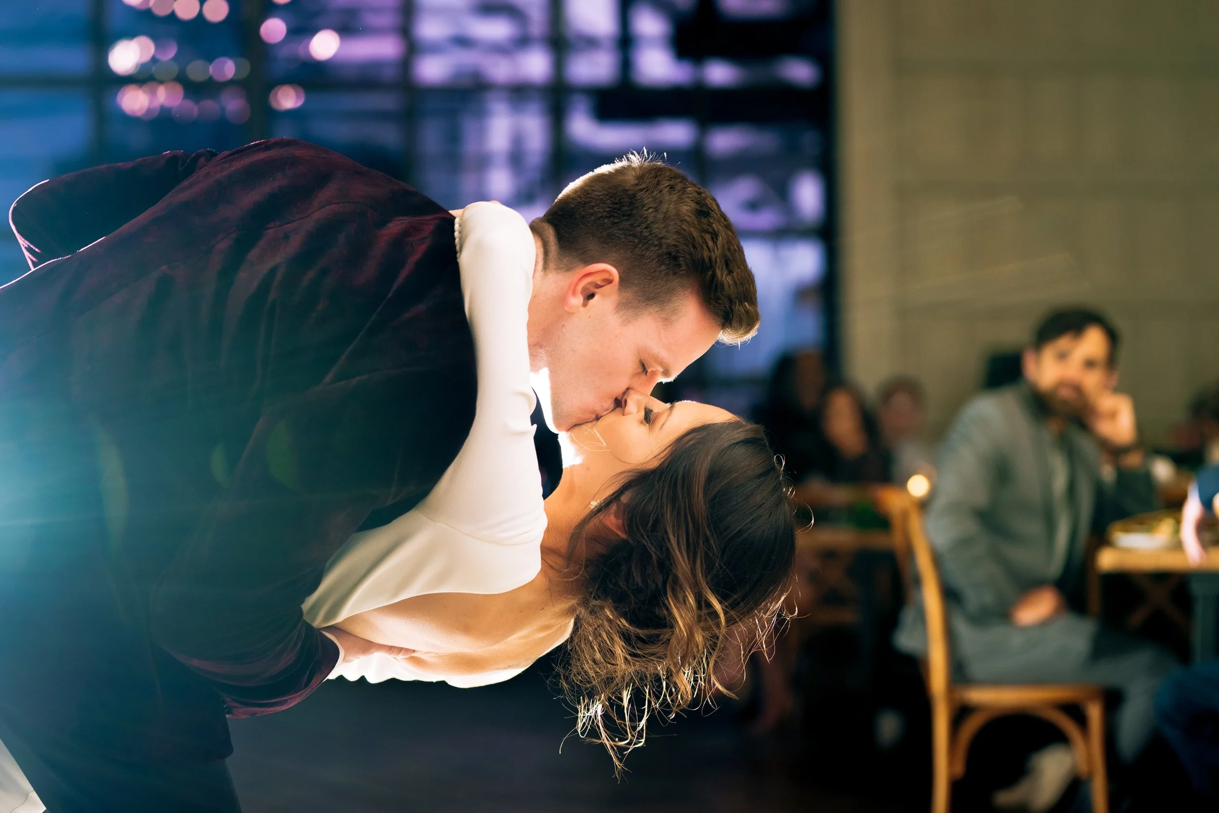 A man and woman are sharing a kiss, with the man leaning over the woman in a dimly lit room, possibly at a celebration or event, with other people sitting at tables at a north Georgia wedding.  Wedding Photography by Sowing Clover Photography.