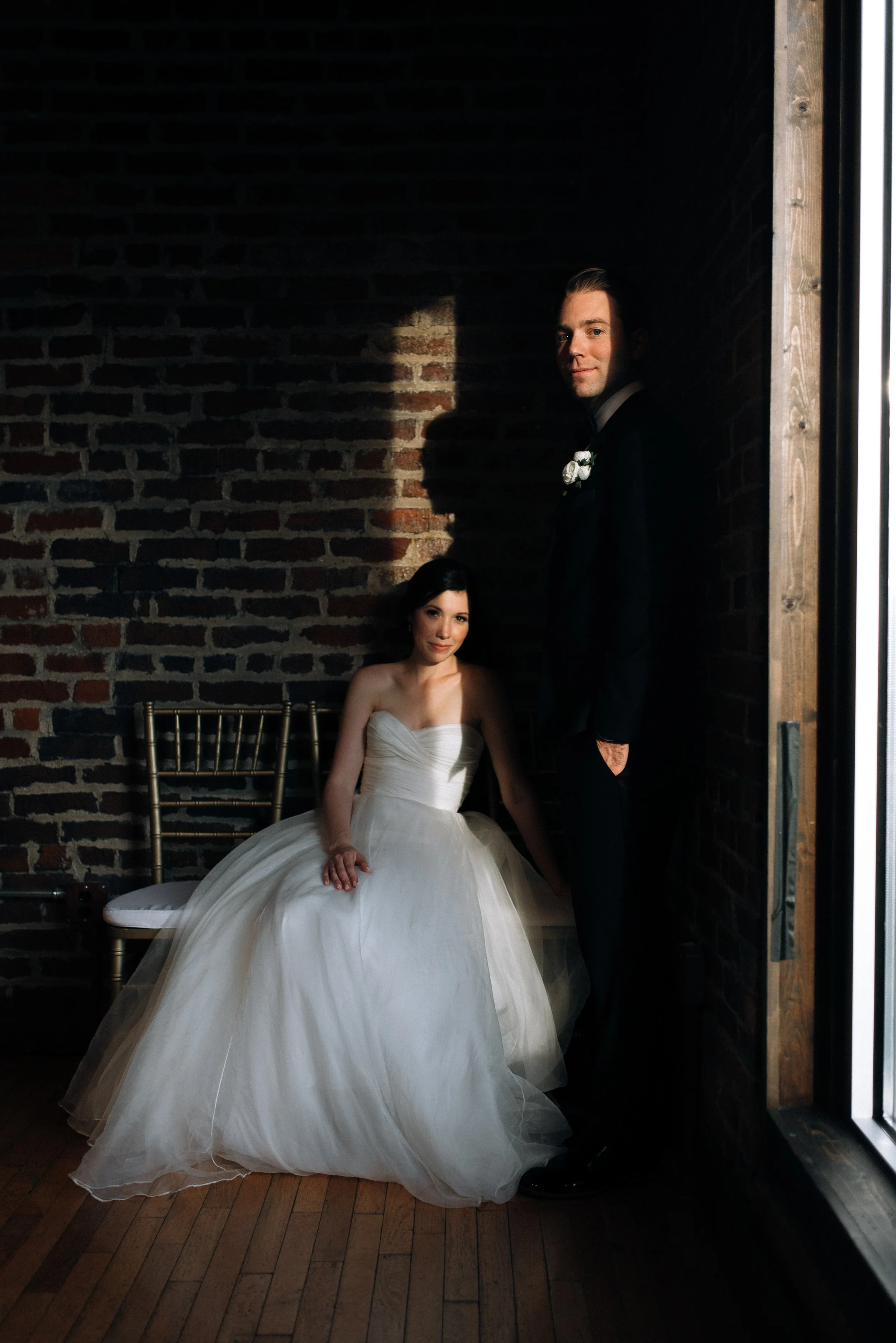 A bride sitting on a chair and a groom standing beside her, both in wedding attire, inside a dimly lit room with a brick wall background downtown Nashville. Wedding photography by Sowing Clover Photography.