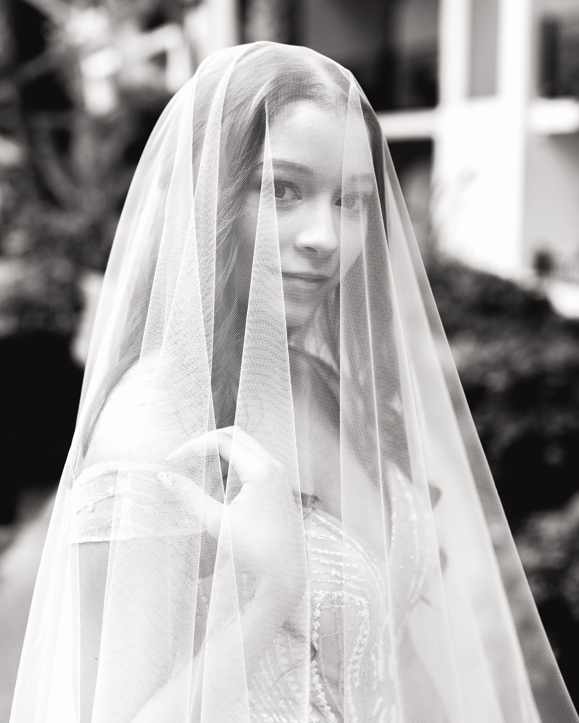Black and white photo of a bride with a veil partially covering her face, smiling, with a blurred outdoor background. Gaylord Opryland Resort. Wedding photography by Sowing Clover Photography.