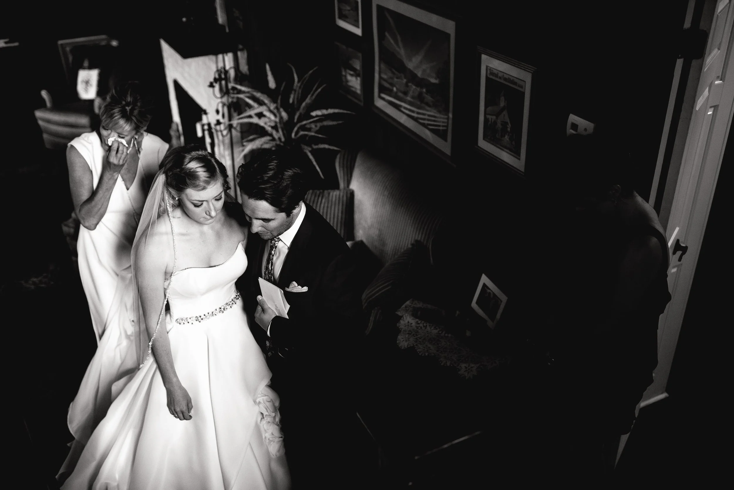 Black and white photo of a bride in a wedding gown with a necklace, and a groom in a suit, reading vows or a speech while a woman wipes away tears in the background. Wedding Photography by Sowing Clover Photography.