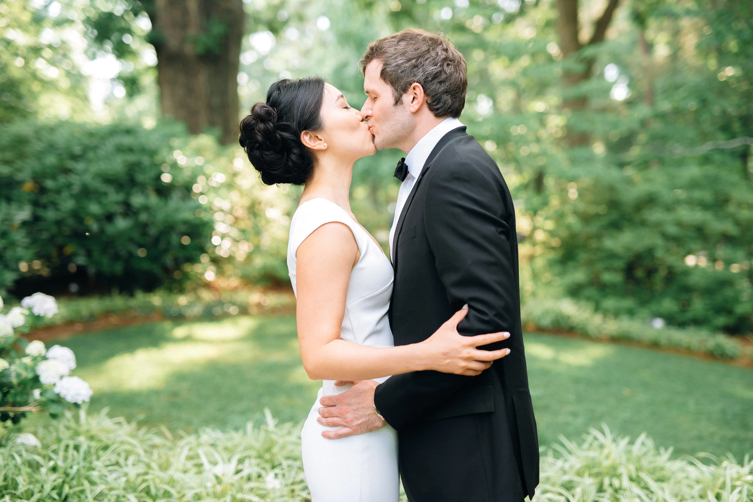 A bride and groom kissing outdoors in a garden during daytime, surrounded by green trees and bushes, with white flowers in the foreground in Atlanta. Wedding Photography by Sowing Clover Photography.