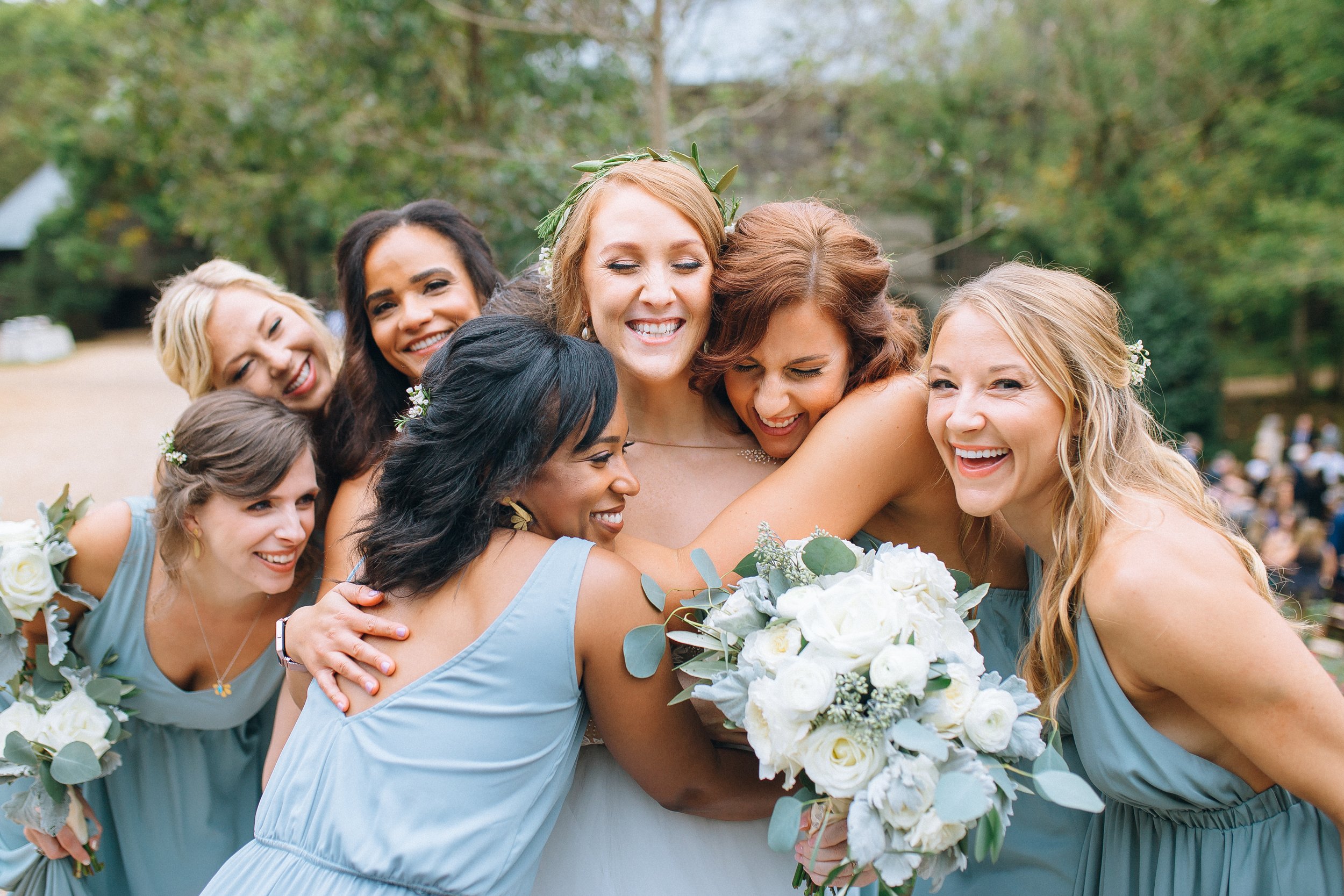 Bridemaids in light blue dresses embracing and smiling at an outdoor wedding celebration, with a bride holding a bouquet of white flowers there and greenery in the background at Yaborough Mill. Wedding Photography by Sowing Clover Photography.