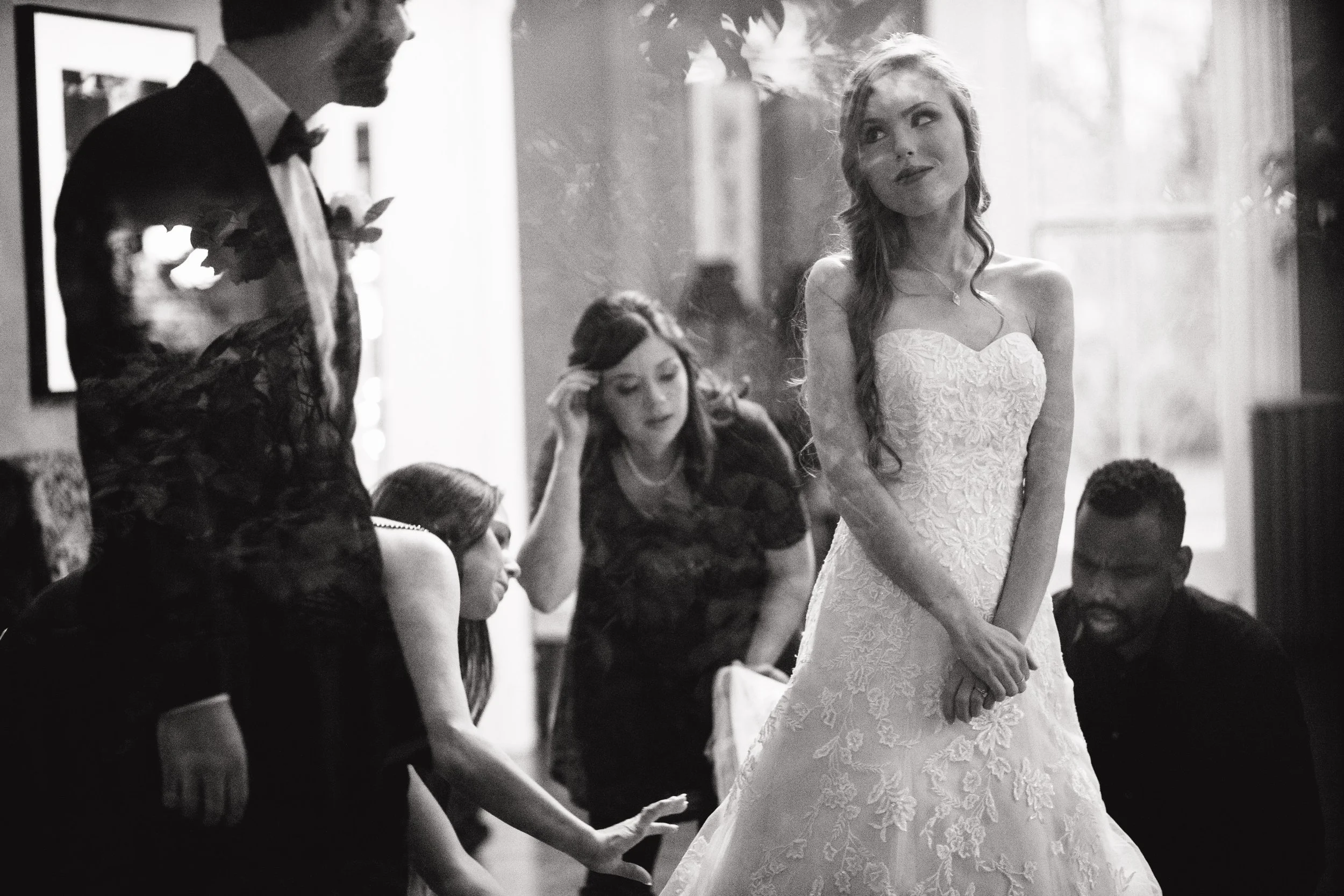 Black and white photo of a wedding reception with a bride in a strapless lace gown standing and smiling, surrounded by seated guests and a man in a tuxedo. Wedding photography by Sowing Clover Photography. Nashville, TN