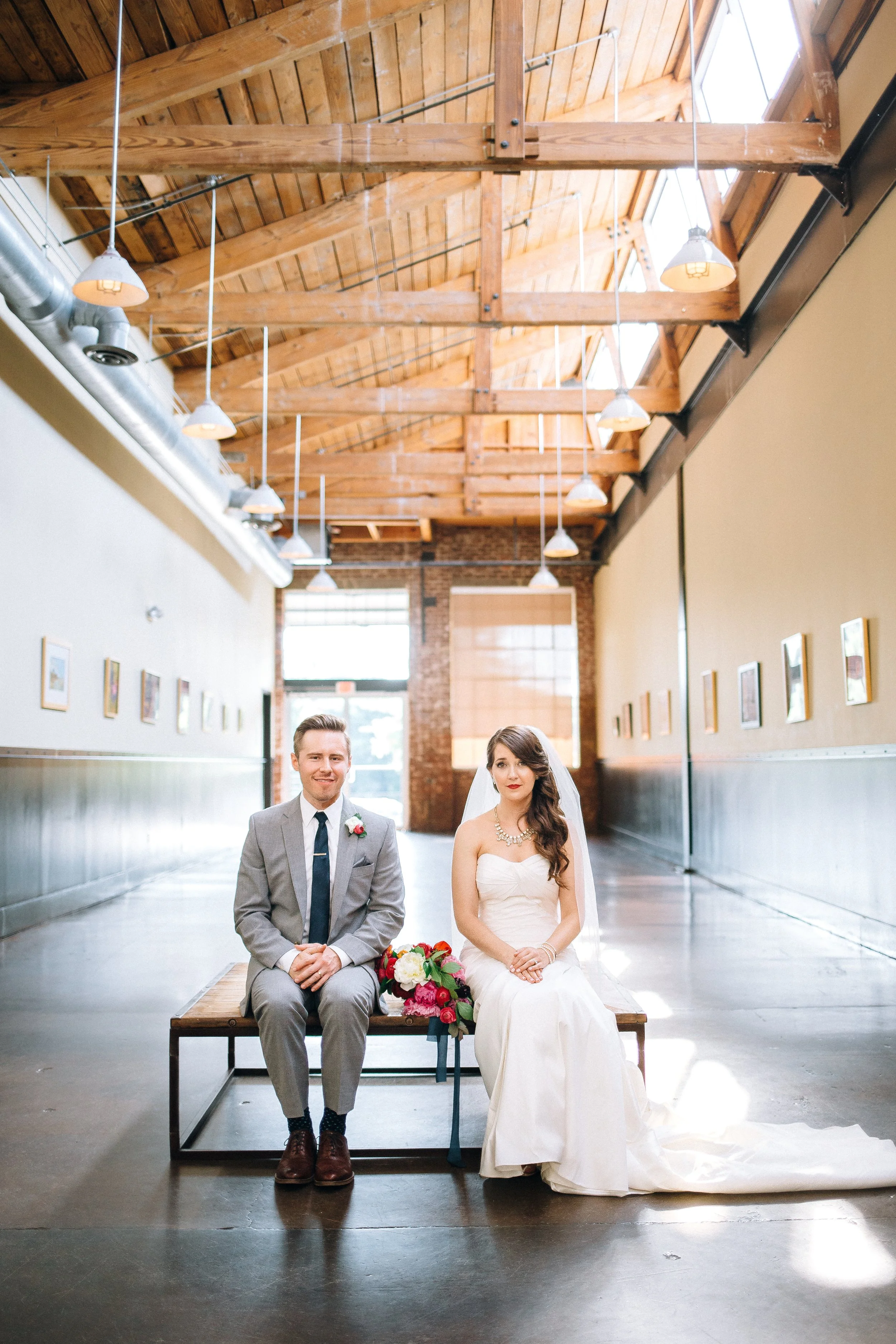 A bride and groom sitting on a wooden bench in an industrial-style venue with a high wooden ceiling, hanging lights, and large windows, during their wedding at the Foundry in Atlanta. Wedding Photography in by Sowing Clover Photography