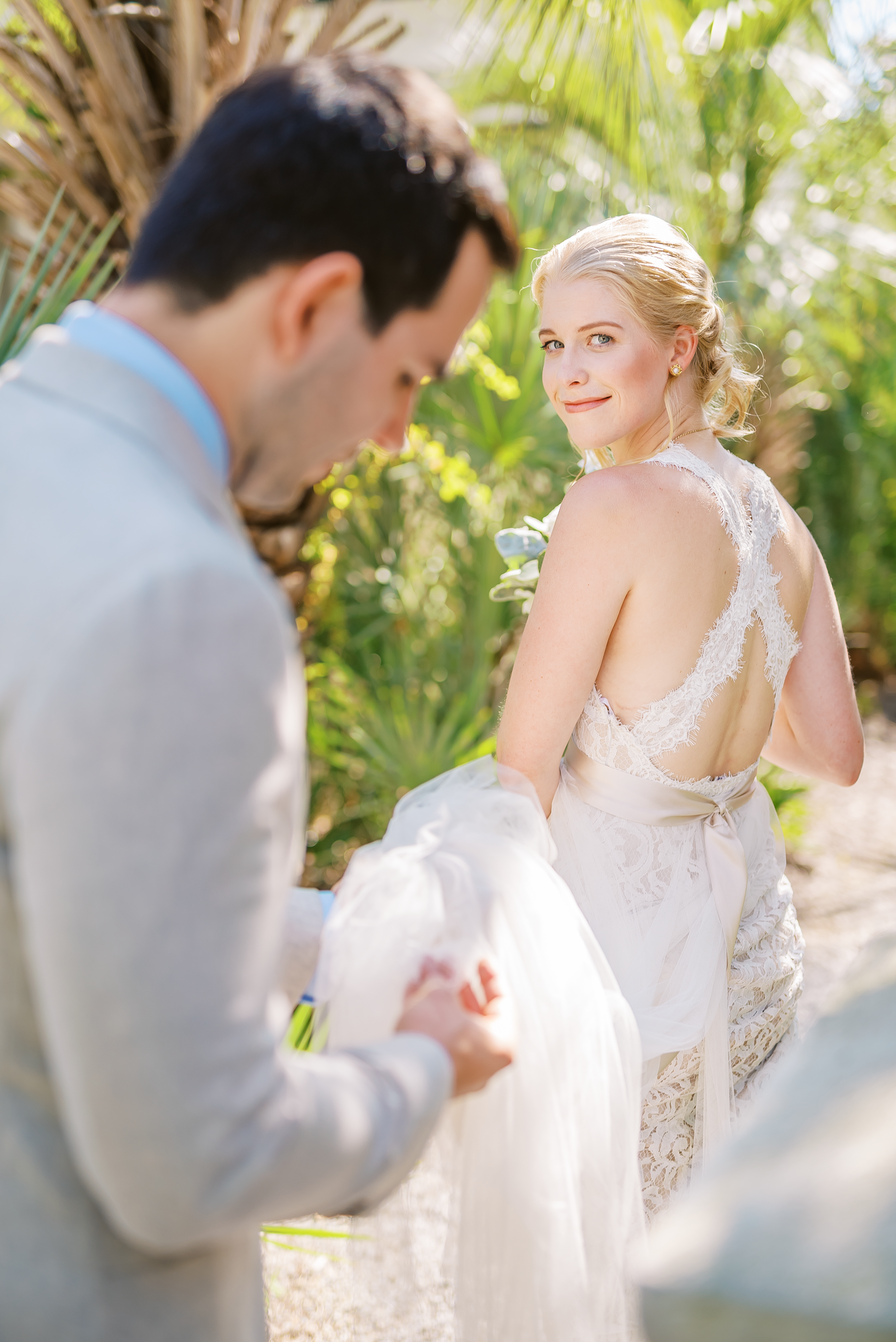Bride in a lace dress with an open back looking over her shoulder at groom holding her train in an outdoor setting with lush green plants.  Florida beach wedding, Wedding photography by Sowing Clover Photography