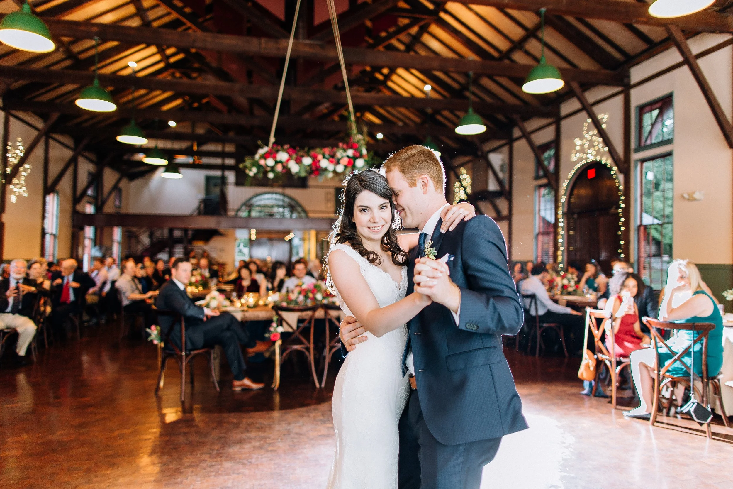 A bride and groom share their first dance at a wedding reception inside a decorated venue with wood beams and hanging floral arrangements. Wedding Photography by Sowing Clover Photography.