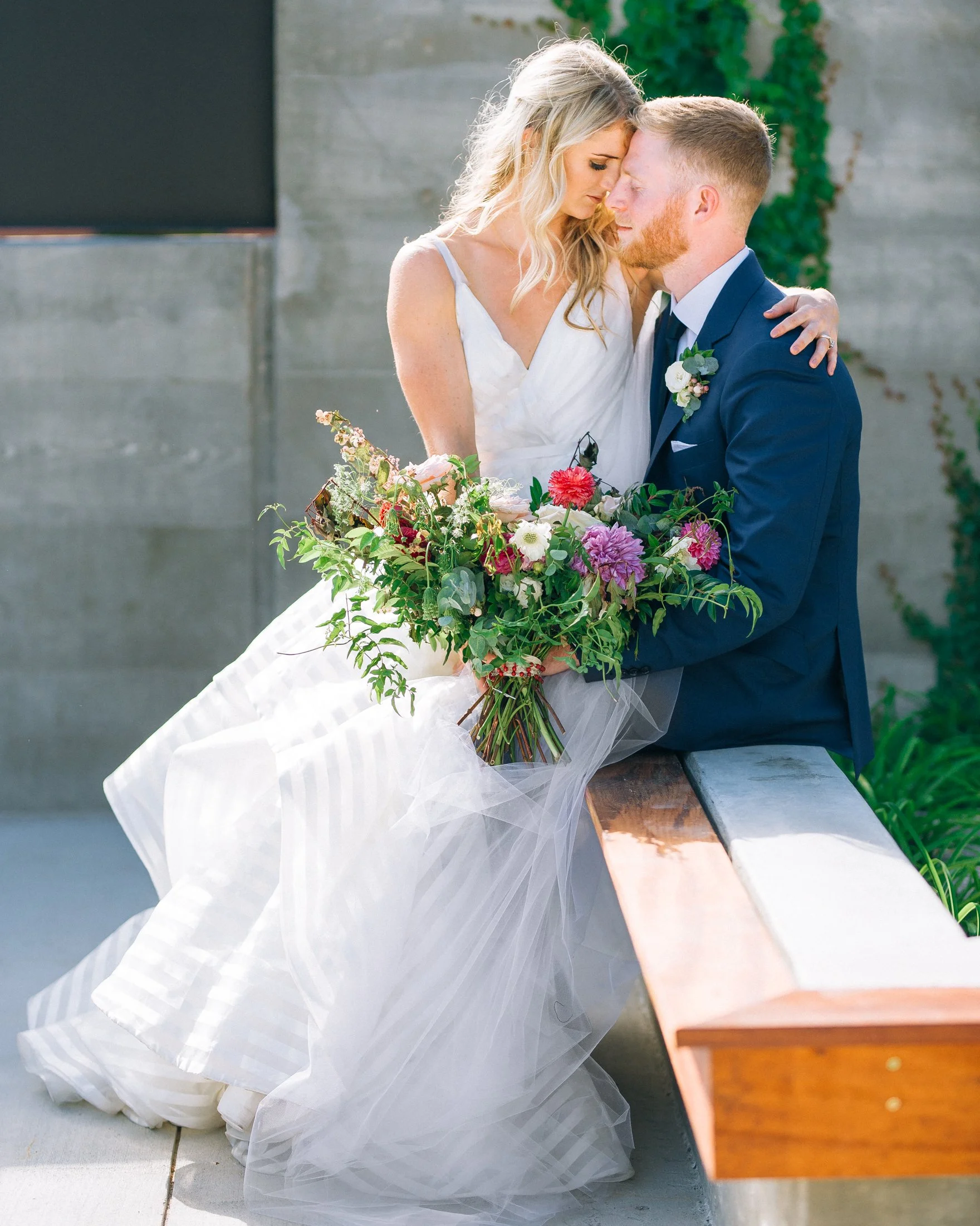 A bride and groom sitting on a bench, with the bride holding a bouquet of colorful flowers. They are leaning their foreheads together with eyes closed at Clementine Hall in Nashville. Wedding Photography by Sowing Clover Photography.
