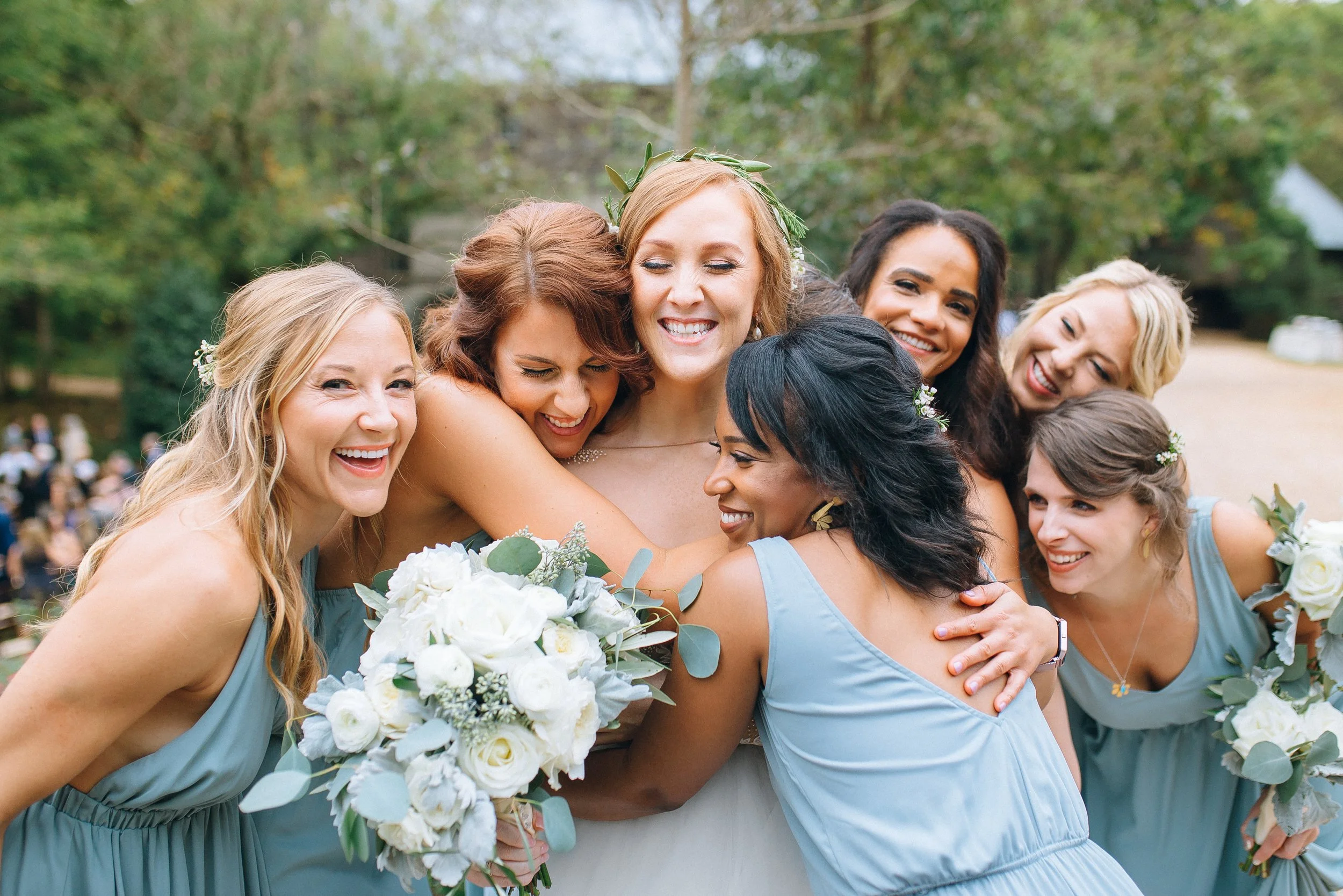 A group of eight women, including a bride, hugging each other outdoors at a wedding, surrounded by greenery at Yarborough Mill. Wedding Photography in by Sowing Clover Photography