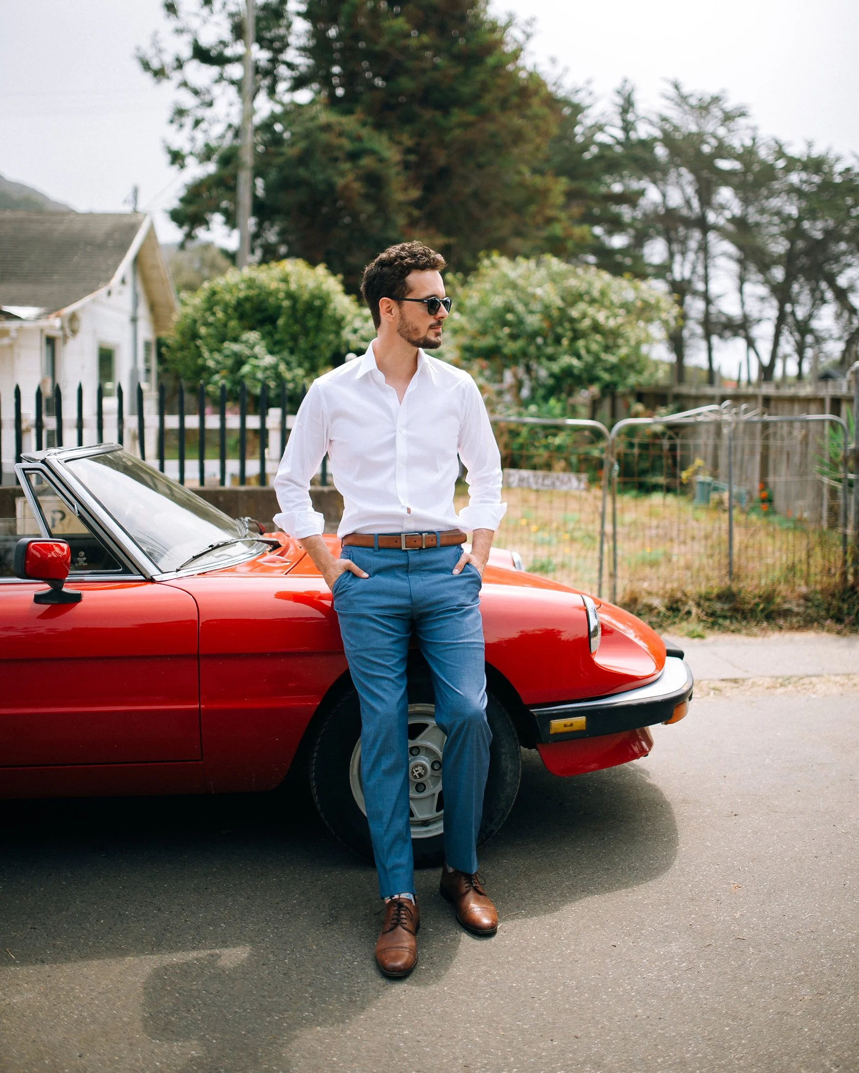 A groom wearing sunglasses, a white button-up shirt, blue pants, and brown shoes, leaning against a red vintage car on a suburban street with trees and houses in the background in San Fransisco. Wedding Photography by Sowing Clover Photography.