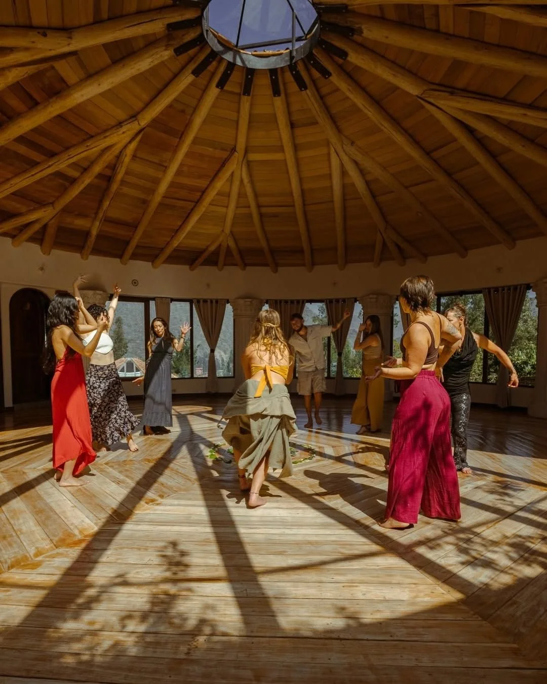 People dancing in a circular formation inside a wooden dome structure with large windows and wooden beams - representing Ginkgo Retreats and community spirit.