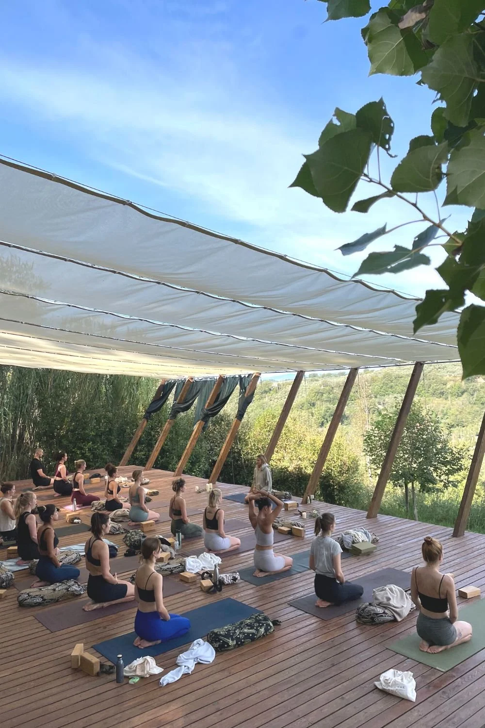 A group of people practicing yoga outdoors on a wooden deck, seated on mats, with a yoga instructor leading them. Surrounding greenery and trees are visible under a blue sky, with partial leaves in the top right corner.