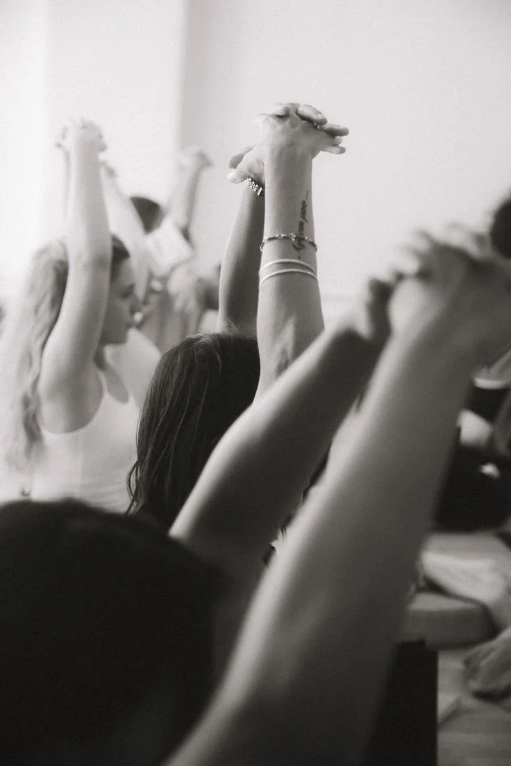 People participating in a yoga or meditation class, sitting with arms raised and hands clasped, inside a bright room.