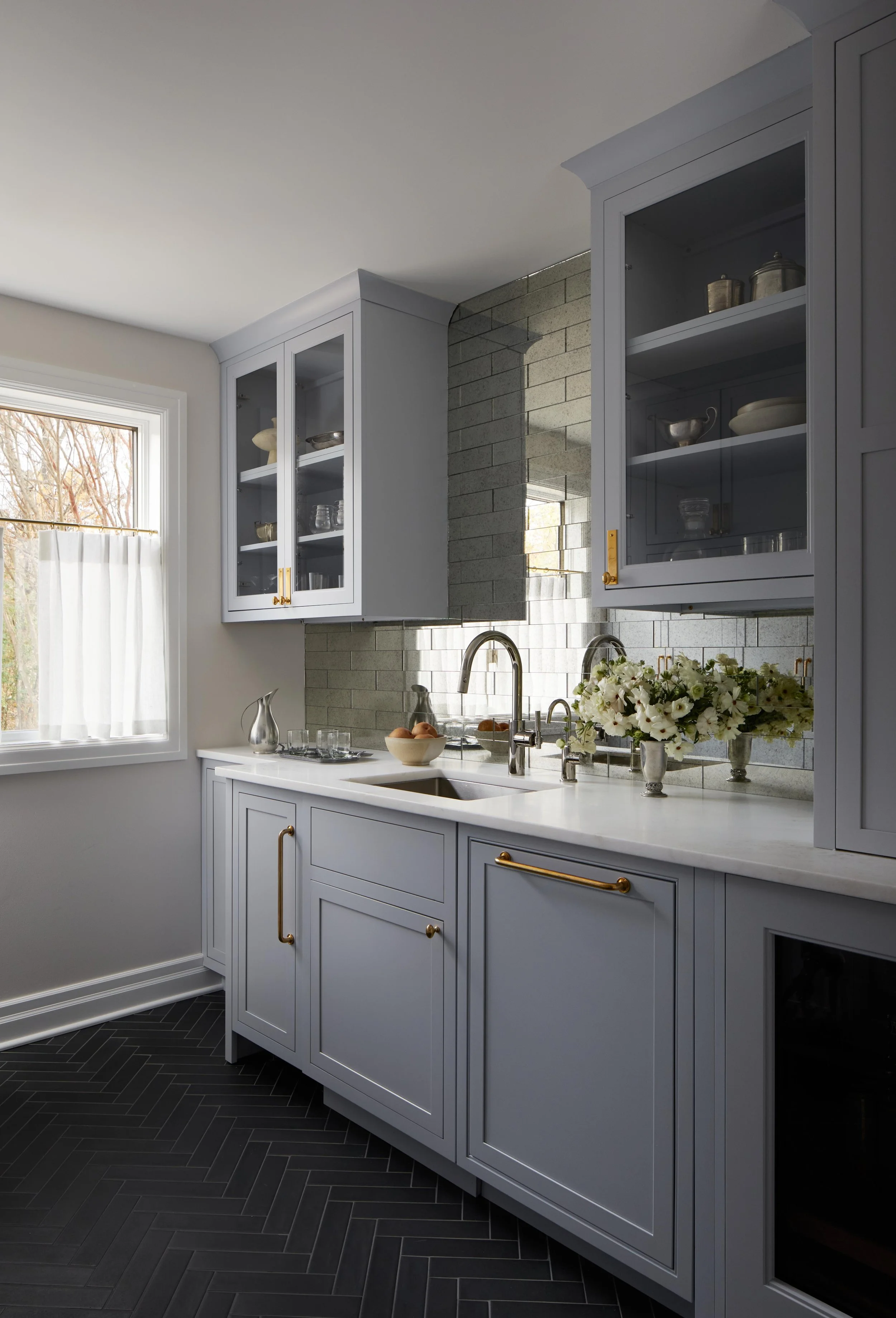 A kitchen with light blue cabinets, a white countertop, a sink with a modern faucet, a window with sheer curtains, and decorative items including a vase of white flowers.