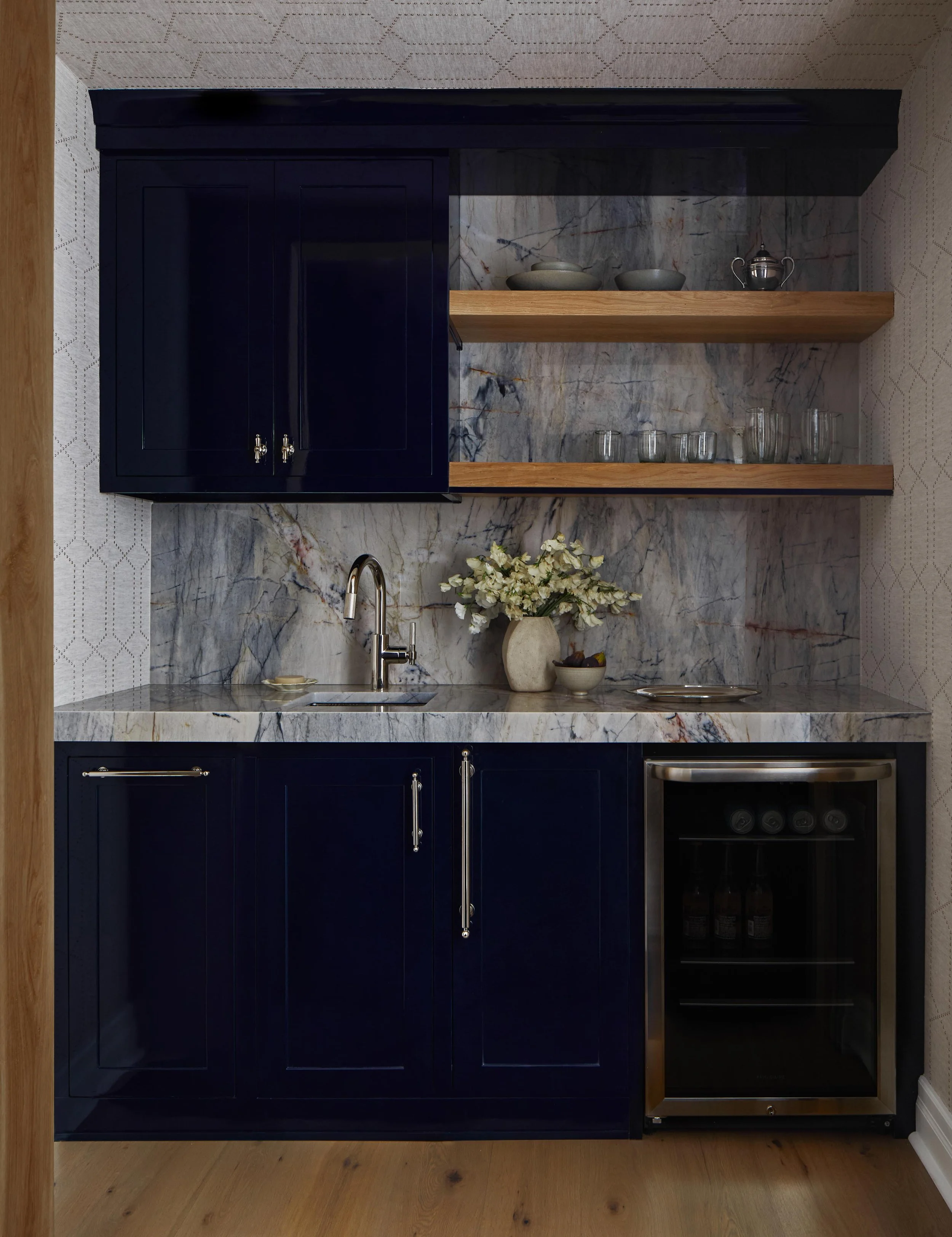 A small modern kitchenette with dark blue cabinets, a marble countertop and backsplash, open wooden shelves with glassware and bowls, a vase with flowers, a small sink with a faucet, and a built-in wine cooler.