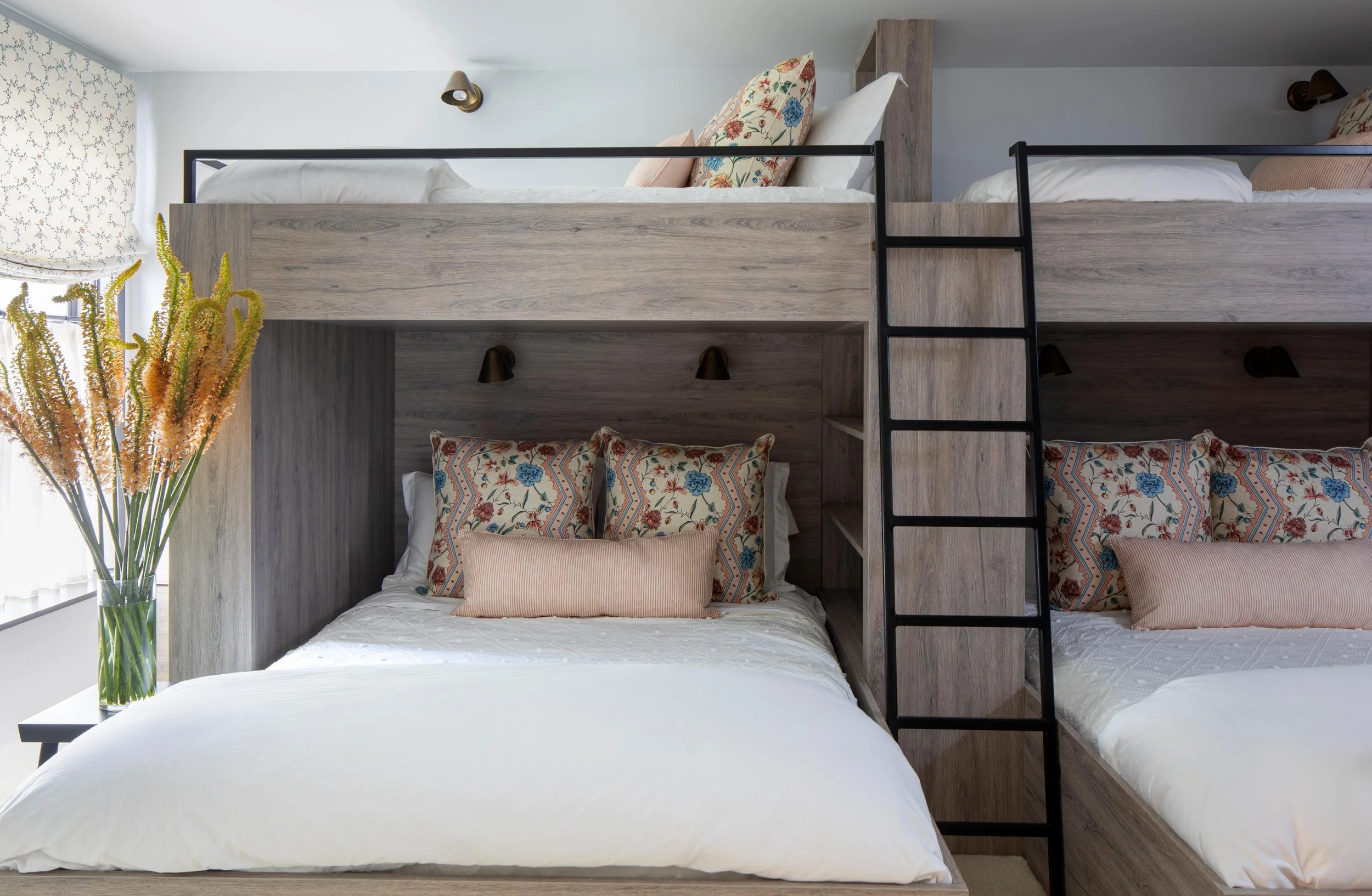 Bunk bed with a double bed below and a single bed above, wrapped in white bedding with floral and striped pillows, located in a bright room with a window and a vase with dried flowers on a side table.
