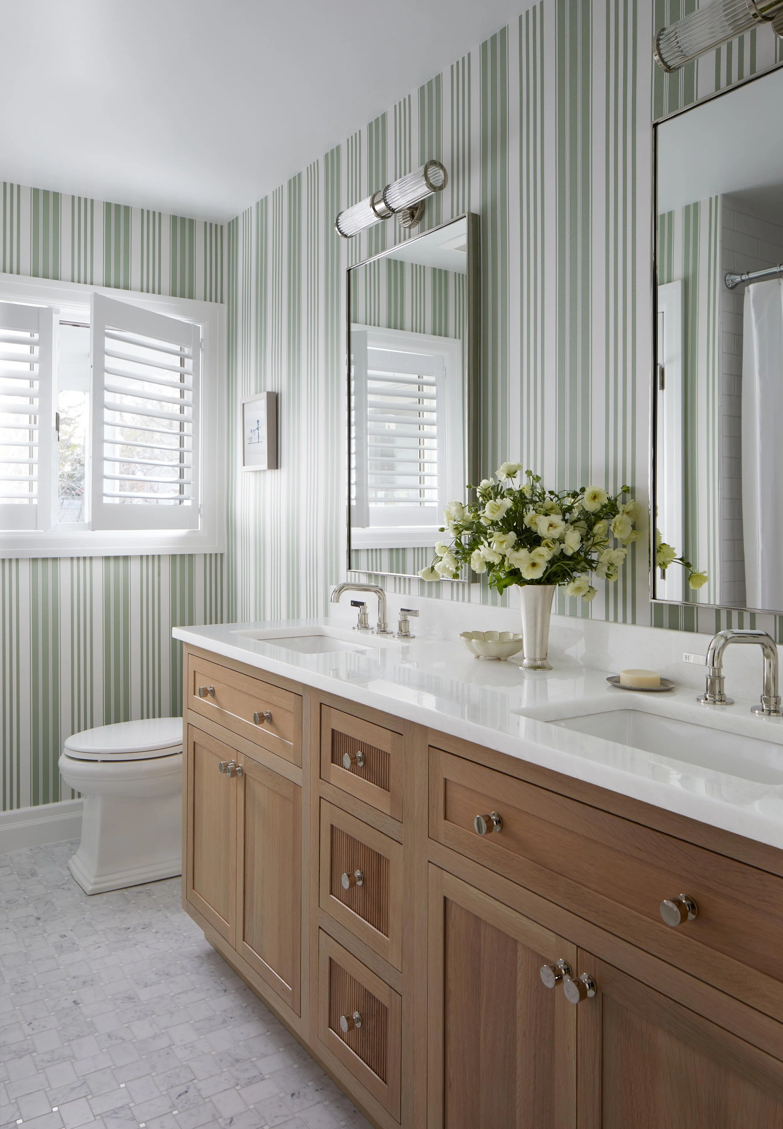 Bathroom with green and white striped wallpaper, a wooden vanity with a marble countertop, a double sink, a large mirror, two wall-mounted lights, a vase of white flowers, and two windows with white shutters.