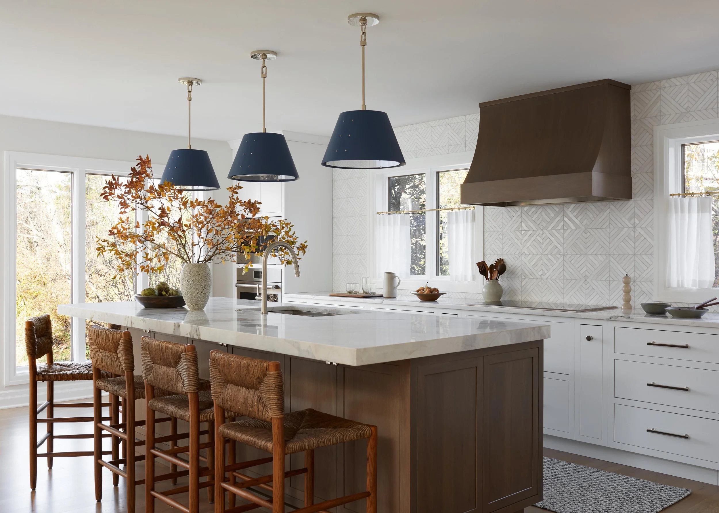 Modern kitchen with white cabinets, a marble island with brown stools, black pendant lights, a range hood, and large windows bringing in natural light.