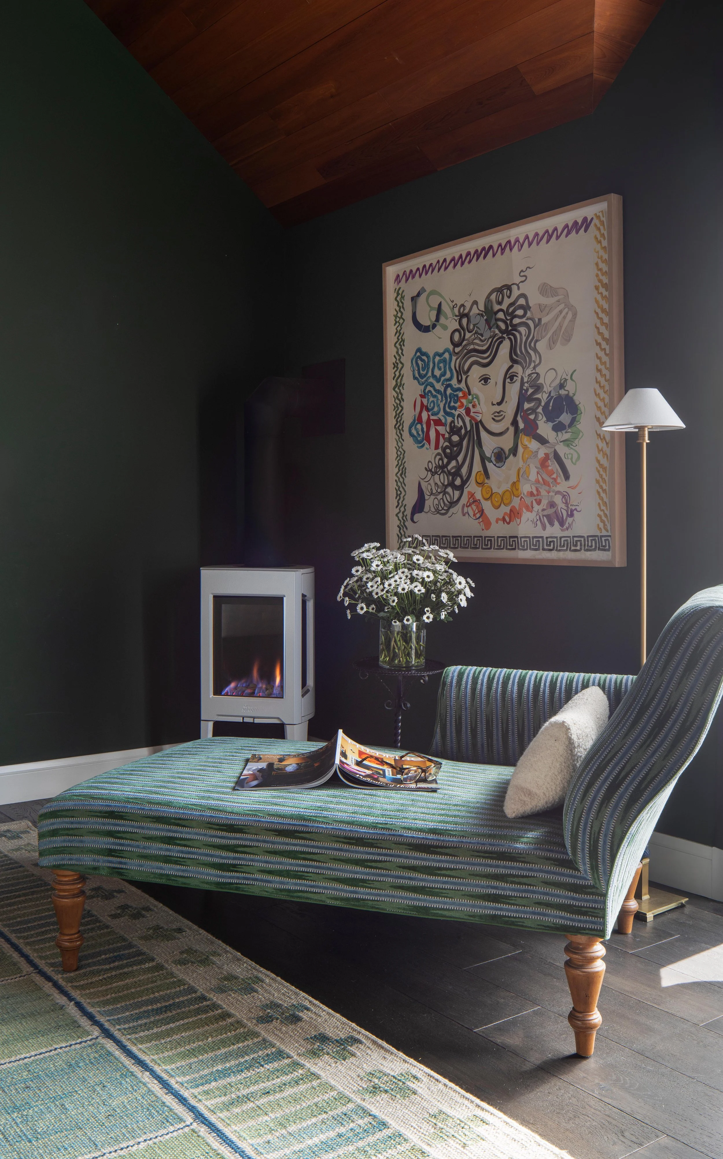 Living room with a chaise lounge, a wall-mounted colorful art piece, a small electric fireplace, a floor lamp, a vase of white daisies on a small table, a magazine on the chaise, and a patterned rug.