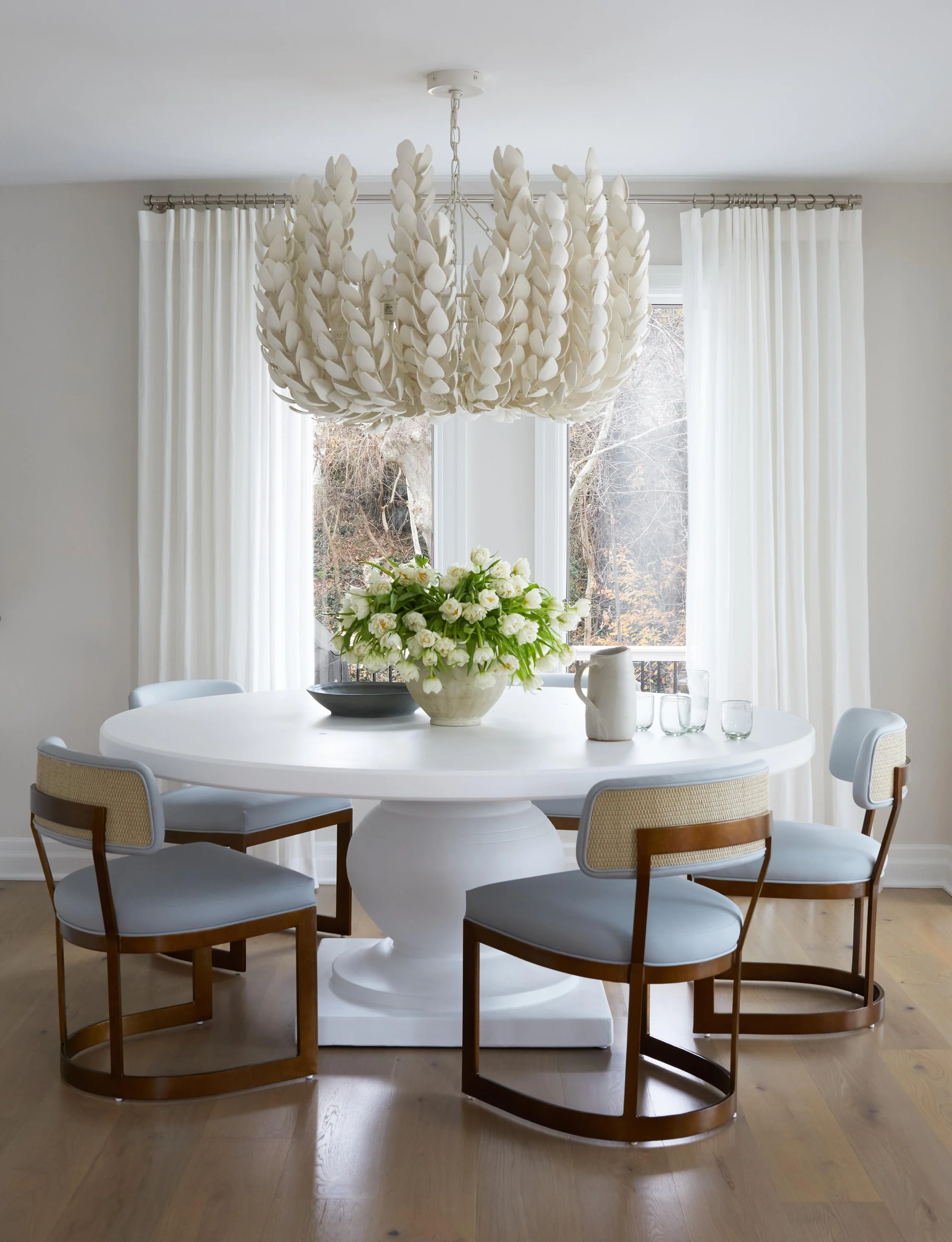 A modern dining room with a white round table, four chairs, a floral centerpiece, and a large statement chandelier in front of a window with white curtains.