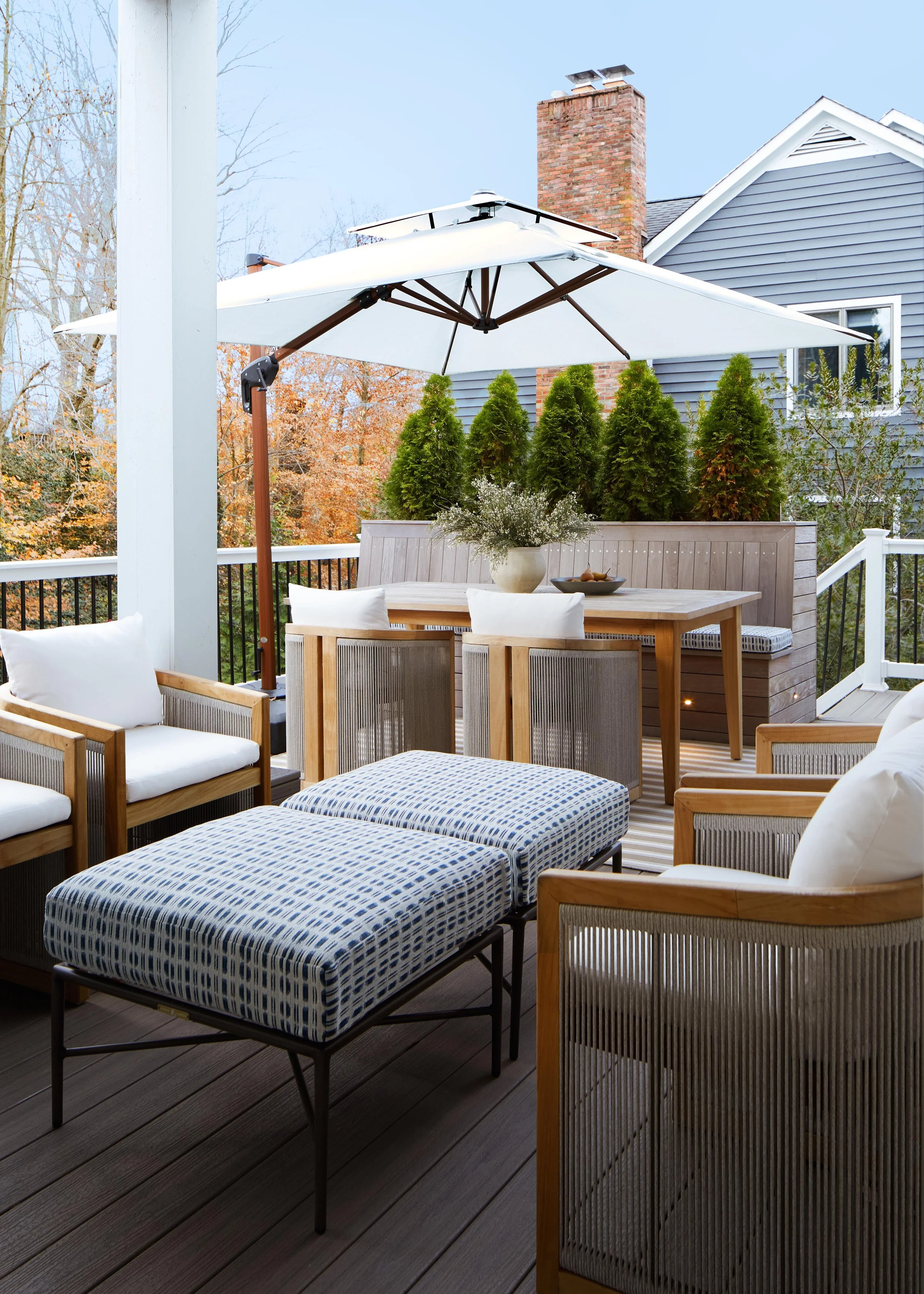 A cozy outdoor patio with wooden furniture, a large white umbrella, potted plants, and a view of trees with fall foliage.