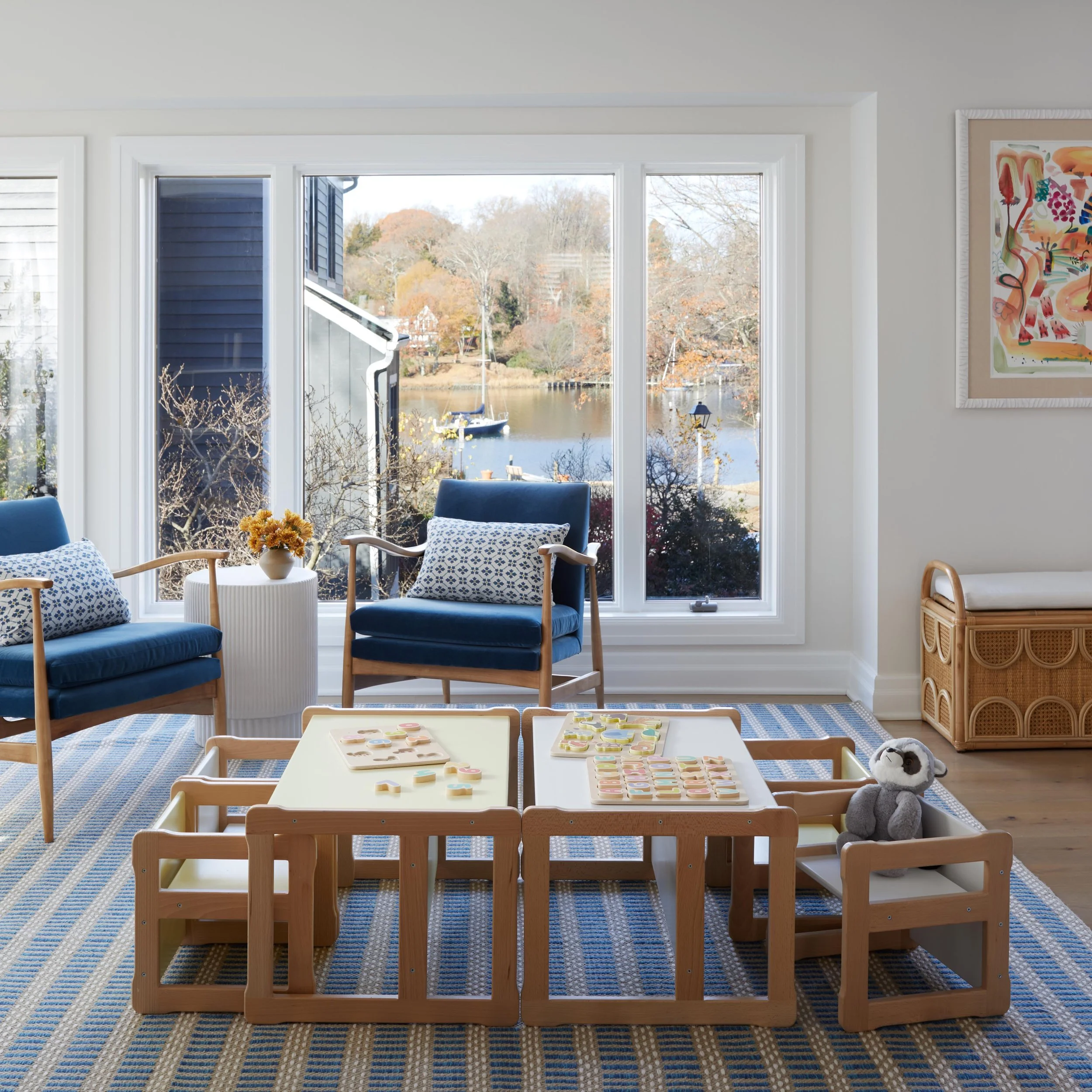 A cozy living room with large windows overlooking a lake and trees. Two blue armchairs with white and blue pillows, a small white table with a vase of yellow flowers, a woven basket, and a children's play table with playdough and toys. A plush gray a