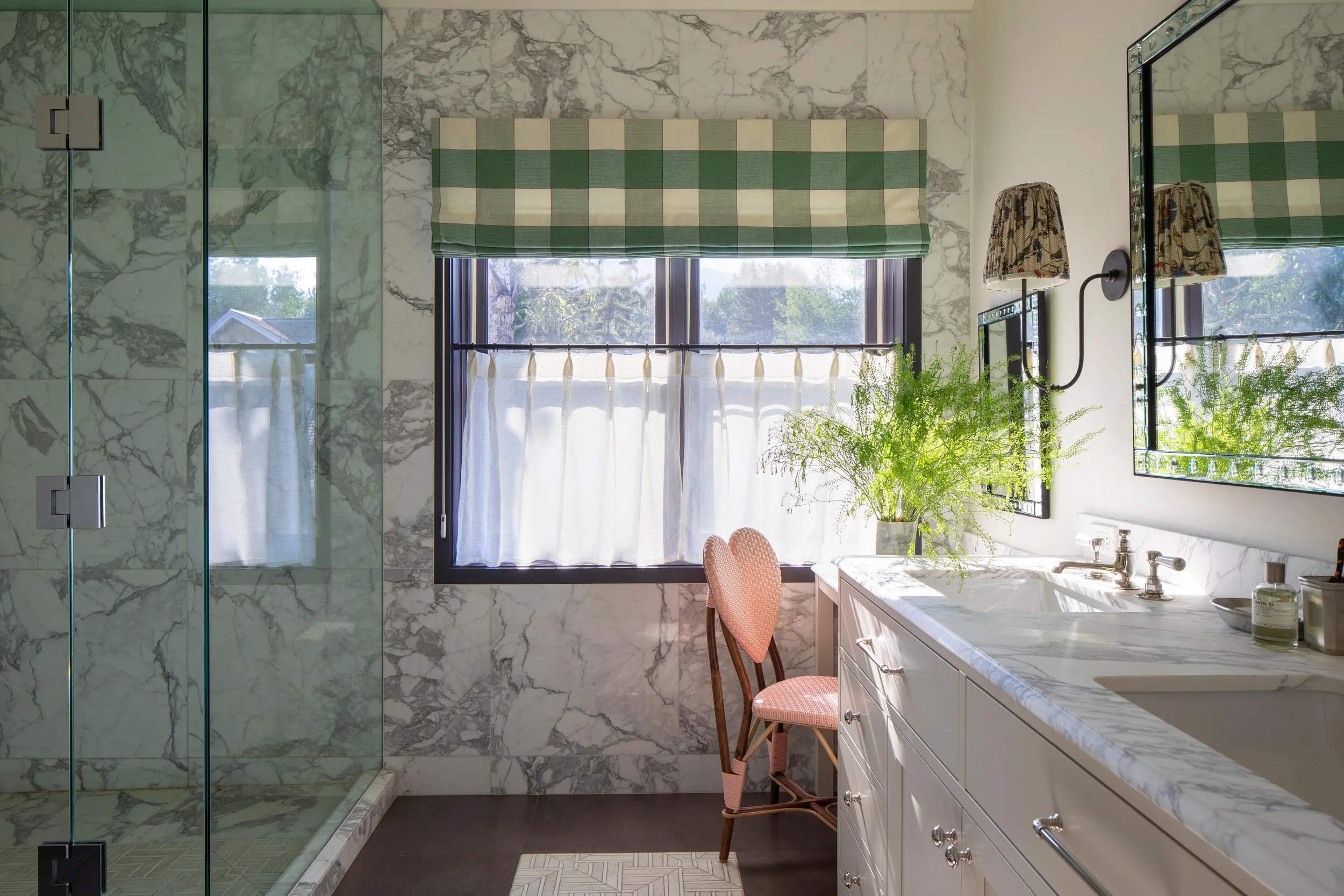 Bathroom with marble walls, a glass shower, a window with a green checkered curtain, a white vanity with a marble countertop, a pink upholstered chair, a potted fern, and a large mirror.