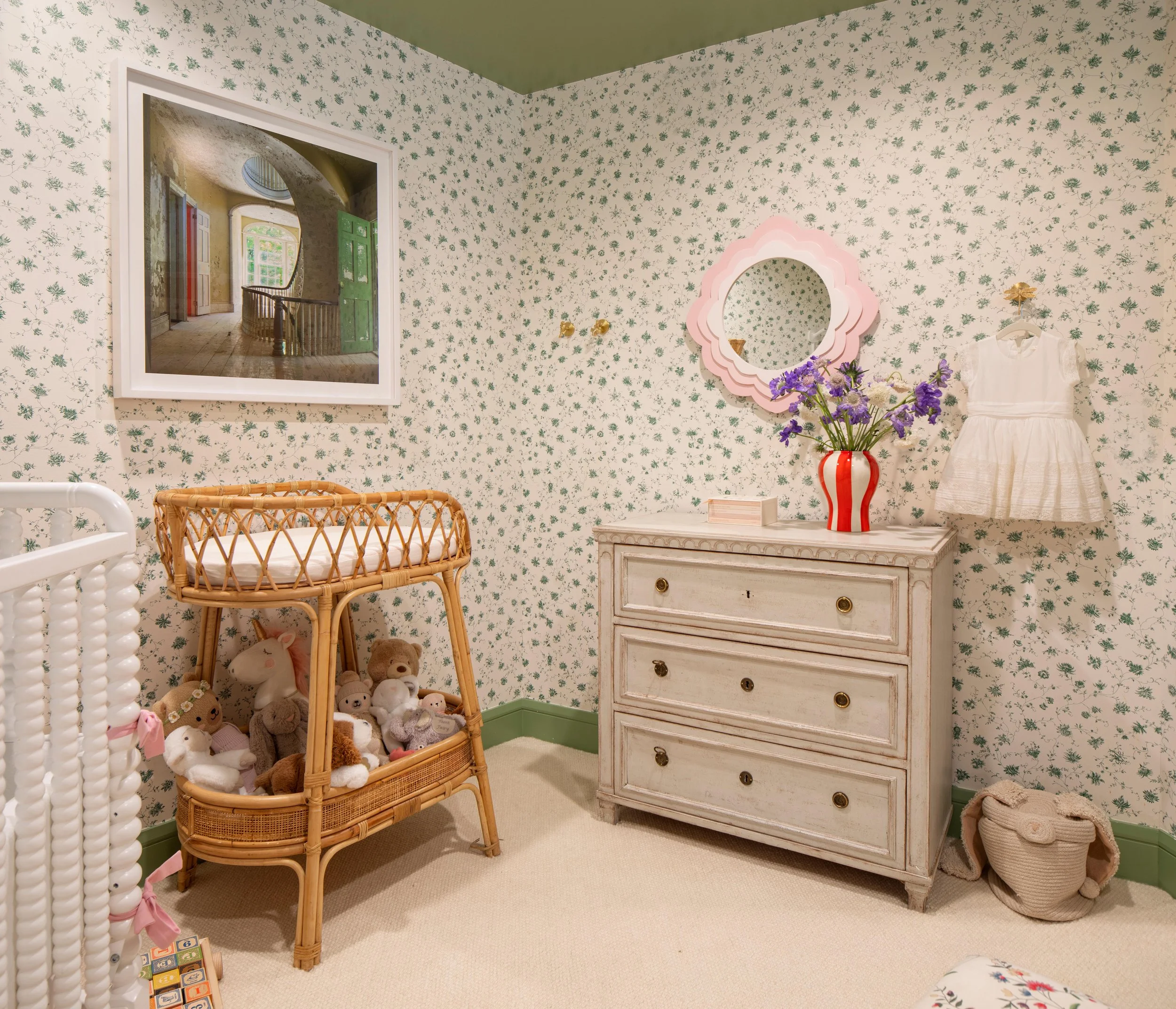 A nursery room with floral wallpaper, a wicker crib filled with stuffed animals, a white dresser with a pink flower mirror above it, a vase with purple flowers, a white dress hanging on the wall, a cream-colored basket on the floor, and a framed pict