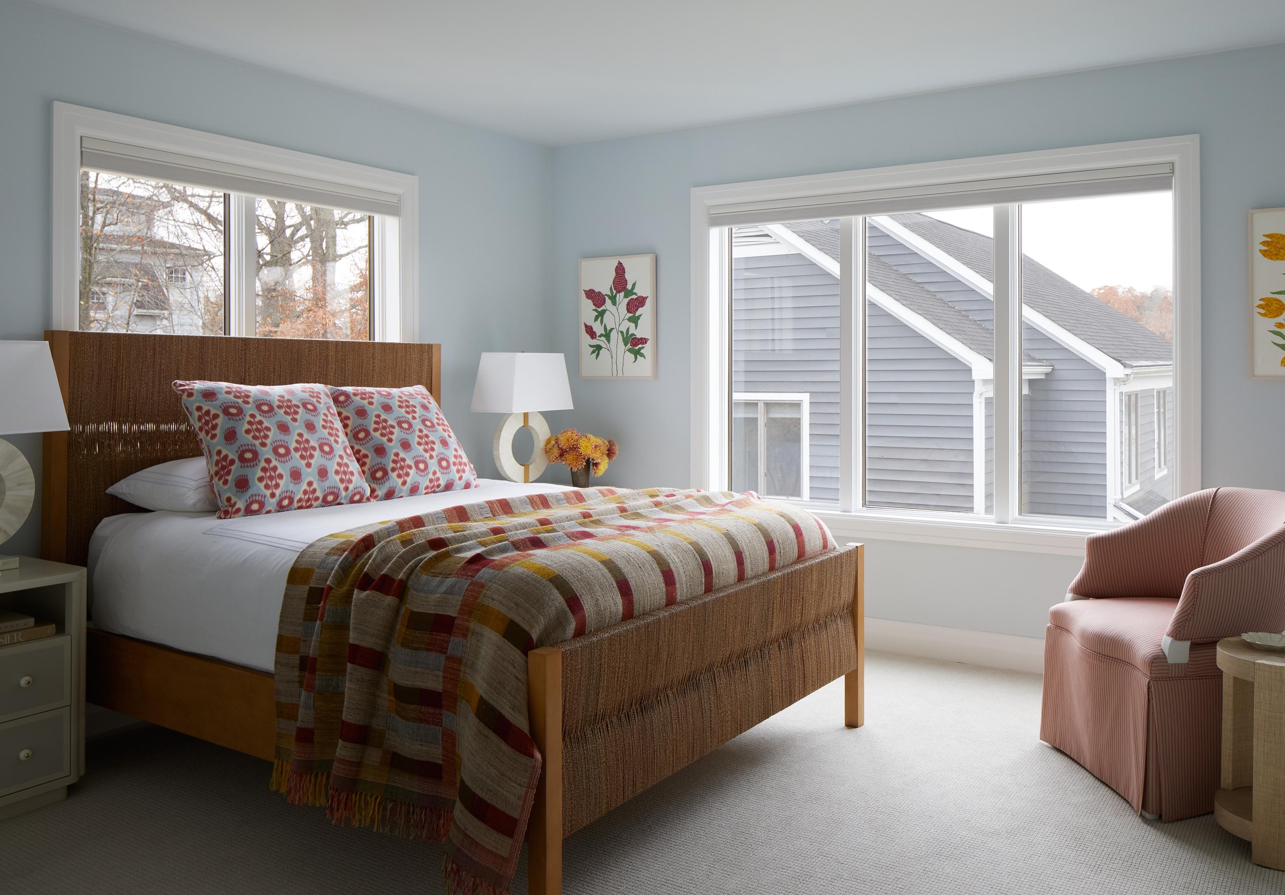 A bedroom with a wooden bed, two pillows with floral patterns, a plaid blanket, two white lamps on nightstands, a pink armchair, and large windows showing neighboring houses.